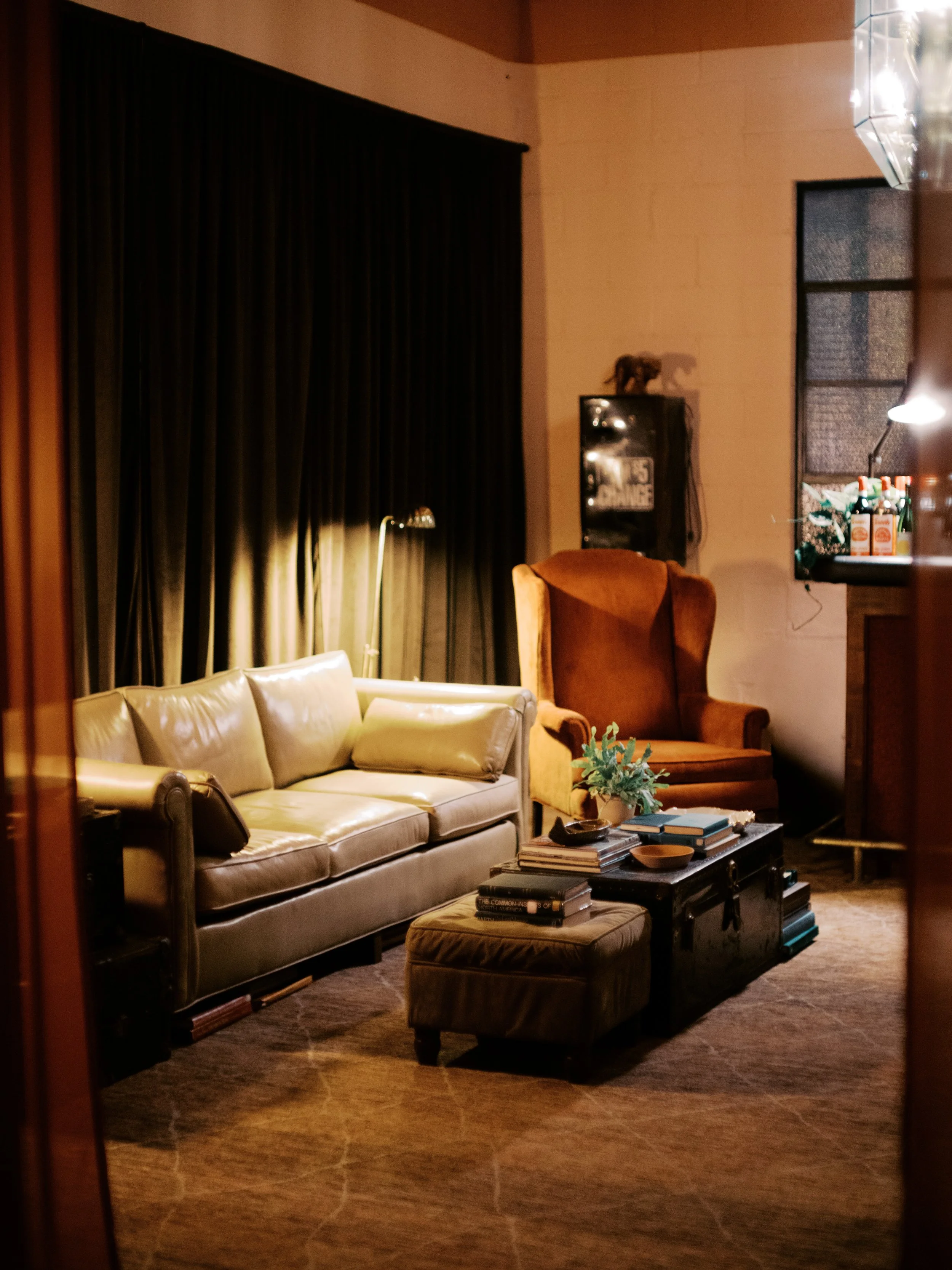 Cozy living room with a beige leather sofa, a brown wingback chair, a dark wooden coffee table with books and a small plant, a black cabinet, and dark curtains.