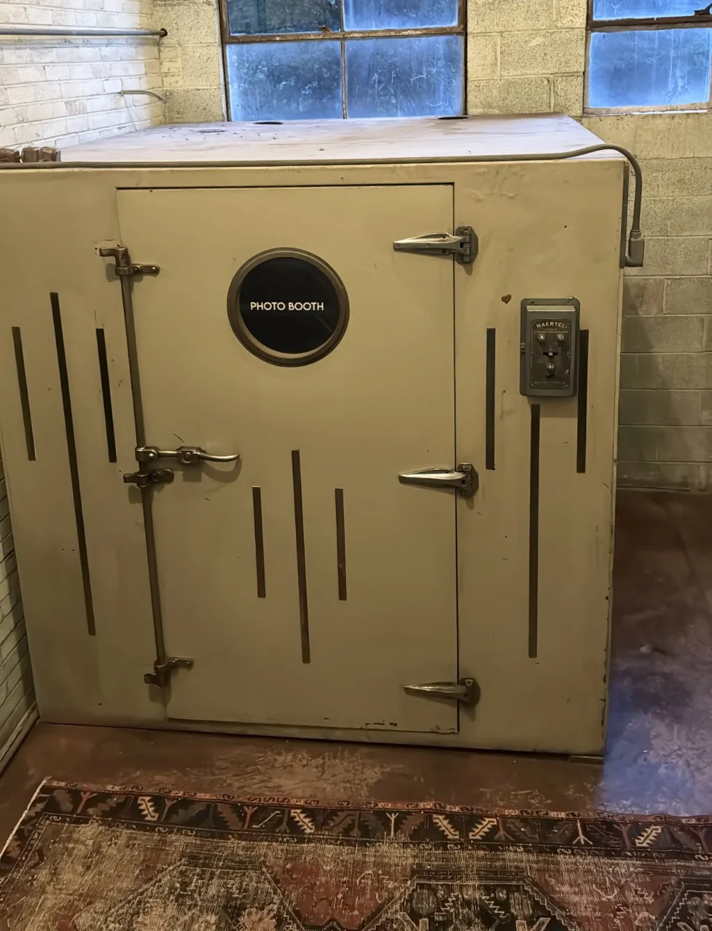An old beige photo booth refrigerator with a small round window labeled 'Photo Booth' and two metal latch locks in a room with brick and concrete walls and blue-tinted windows.
