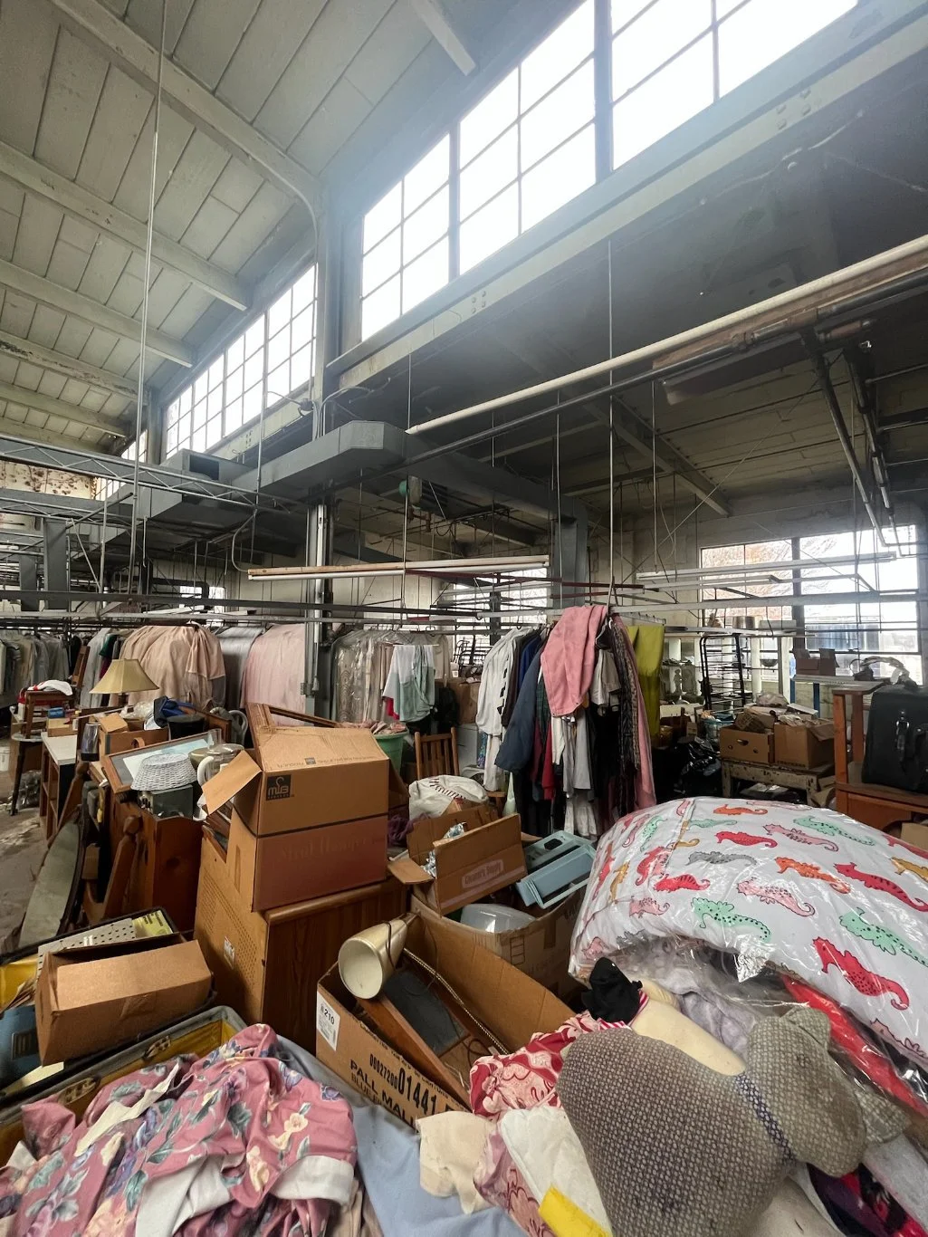 Cluttered indoor space with boxes, clothes, and household items, possibly a thrift store or storage room.