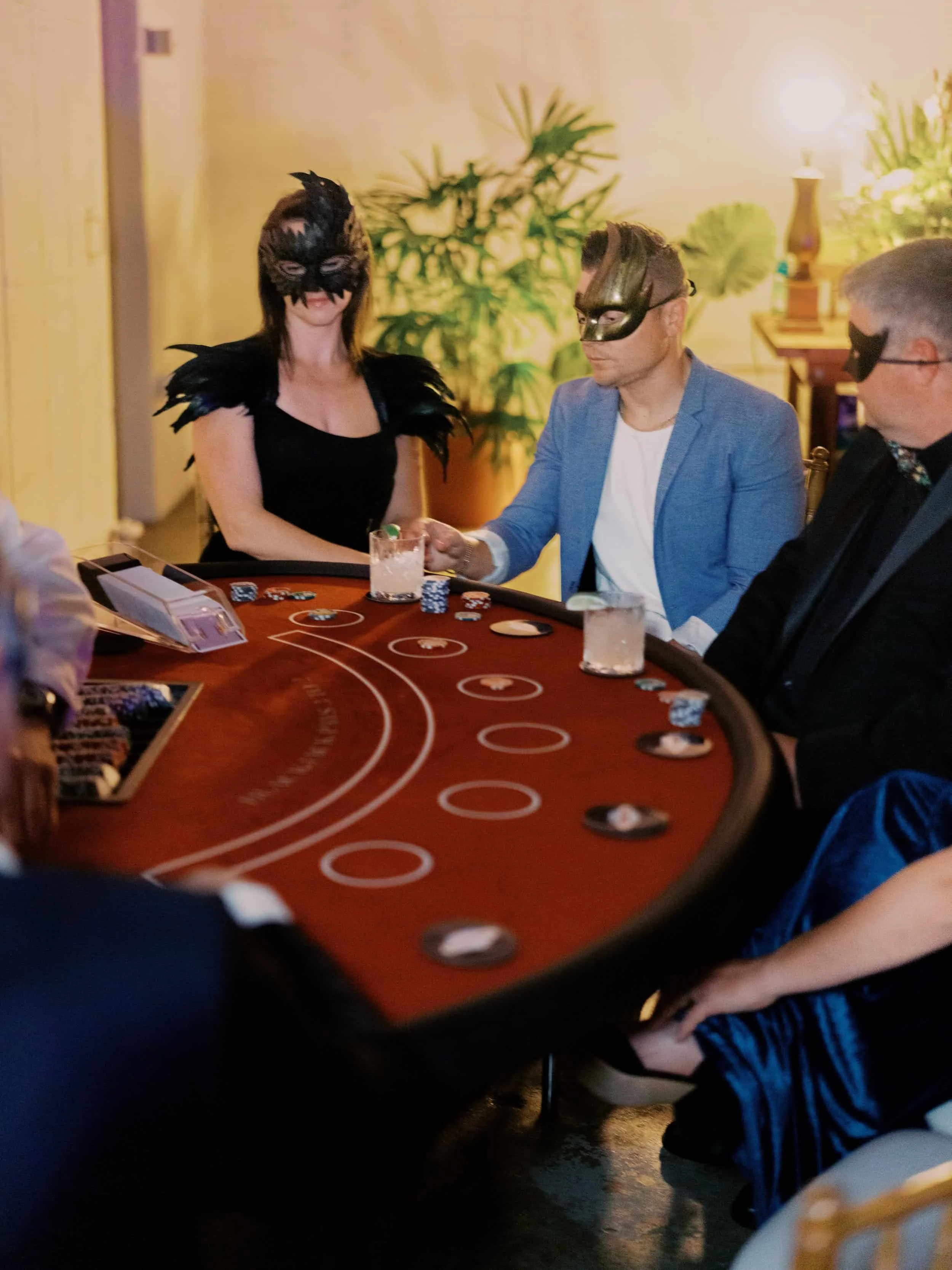 People wearing masks playing poker at a casino table, with drinks and poker chips.