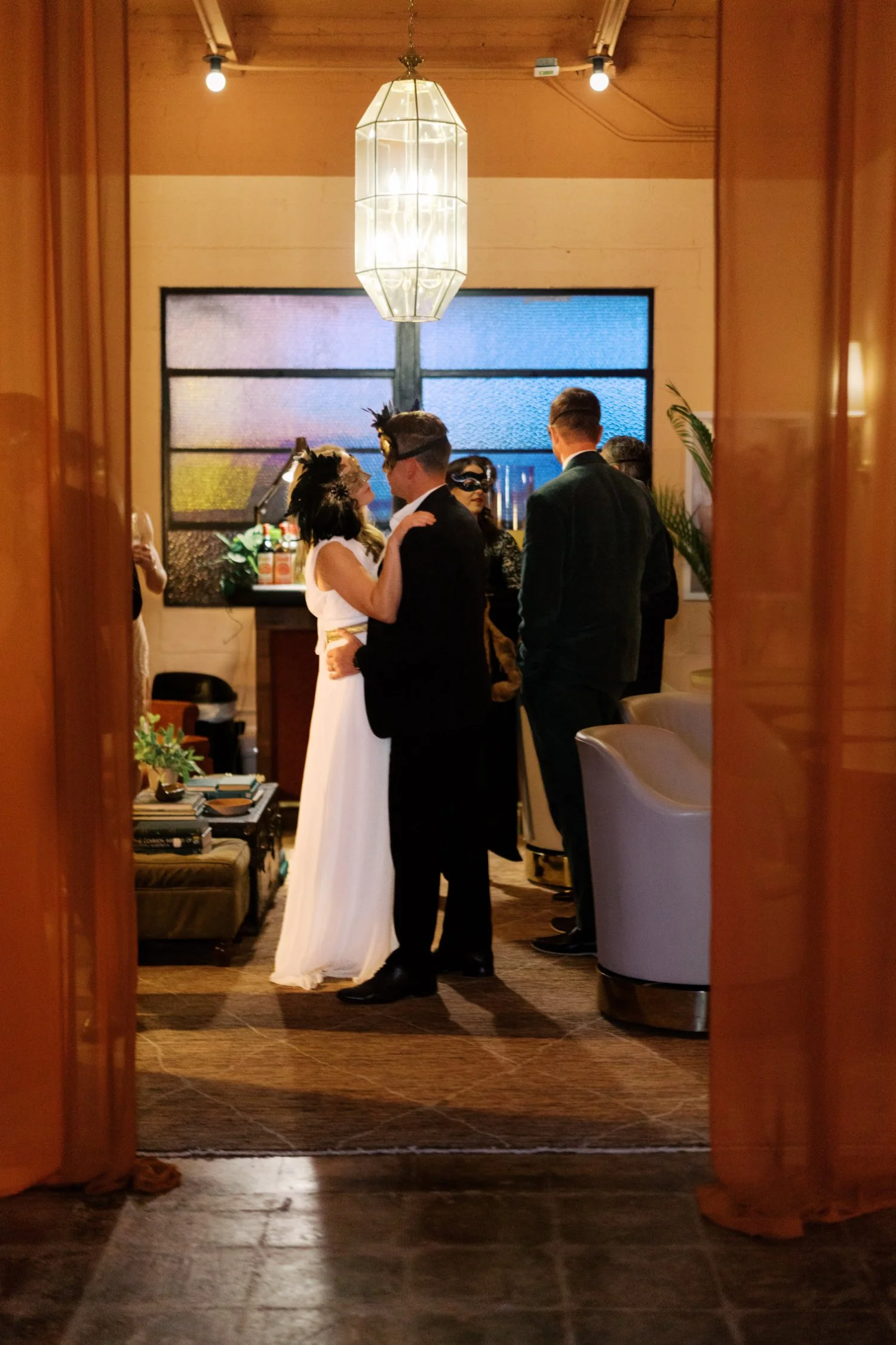 A couple dressed in wedding attire dancing and kissing at a reception, viewed through an open doorway with other guests in the background.