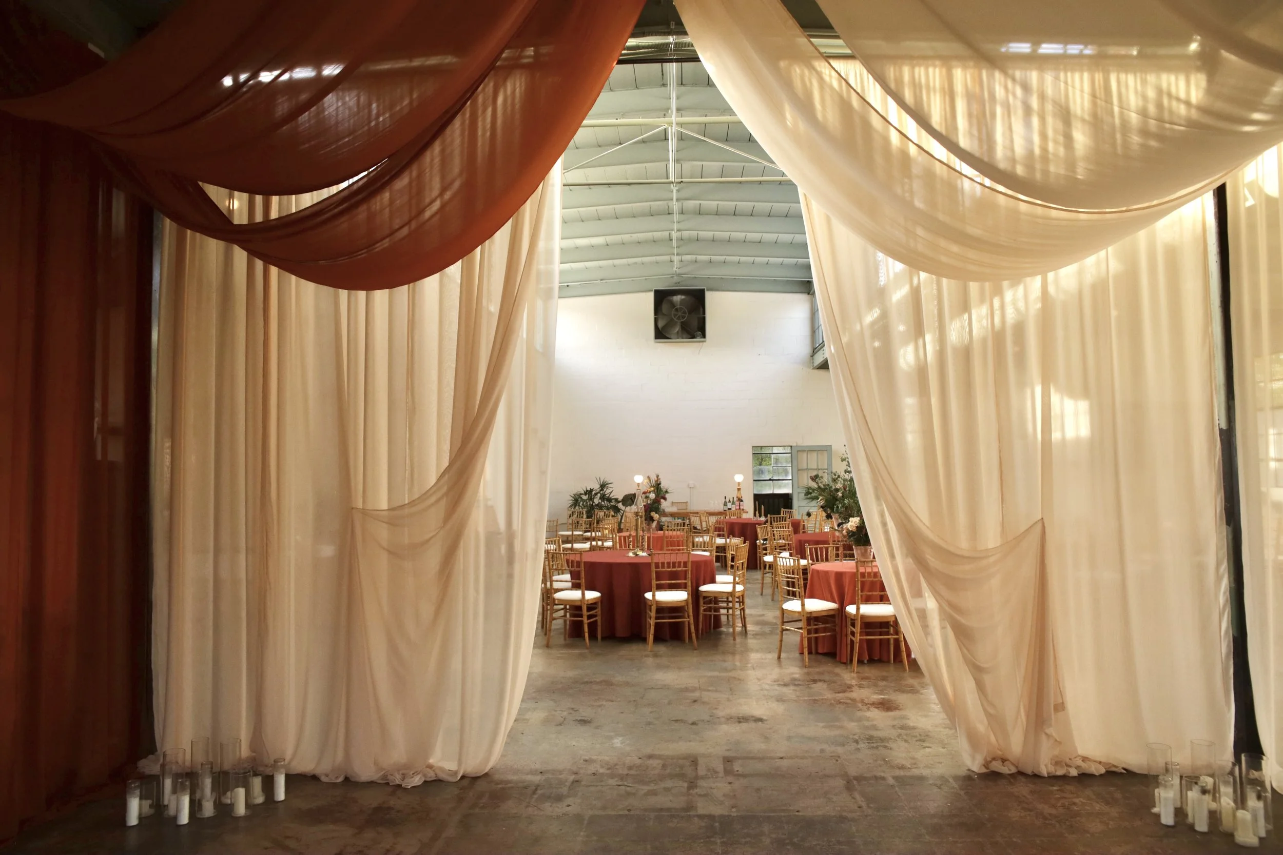 Wedding reception hall with round tables, red tablecloths, gold chairs, and floral centerpieces. Soft draped curtains frame the entrance, with candles on the floor.