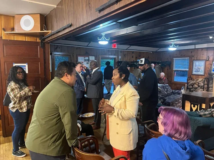 People talking and socializing inside a wooden room with vintage decor and woodpile, featuring both standing and seated individuals, some in conversation and others waiting. Bernice Cooper running for City Council West District Brooklyn Park