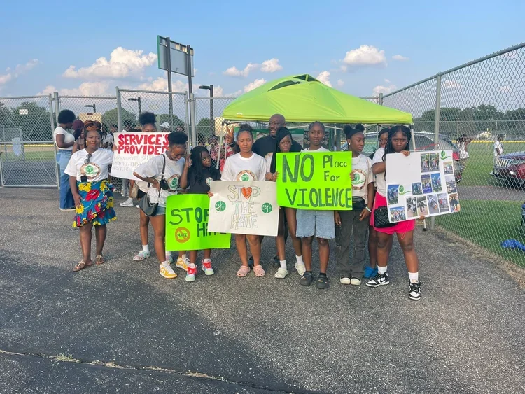 Group of children and adults, Bernice Cooper, holding protest signs against violence, standing near a chain-link fence on a sunny day. Bernice Cooper running for City Council West District Brooklyn Park Minnesota Hennepin County and her family.