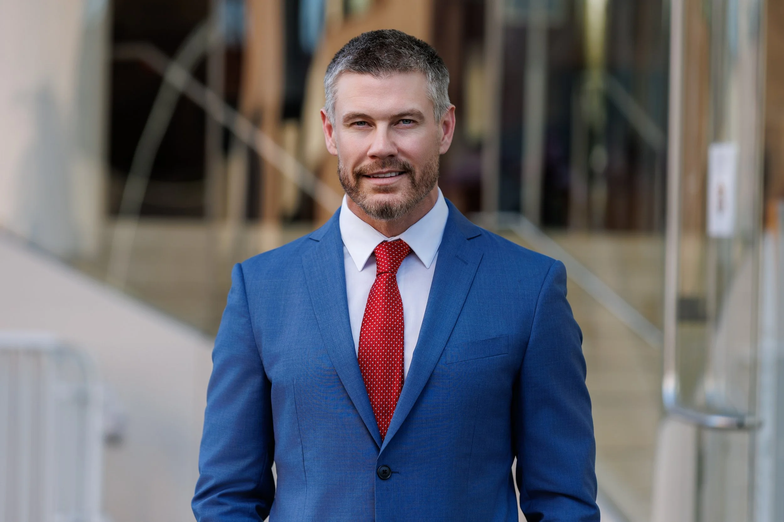 A man with gray hair and a beard wearing a gray suit and a light pink collared shirt, smiling against a dark blue background.