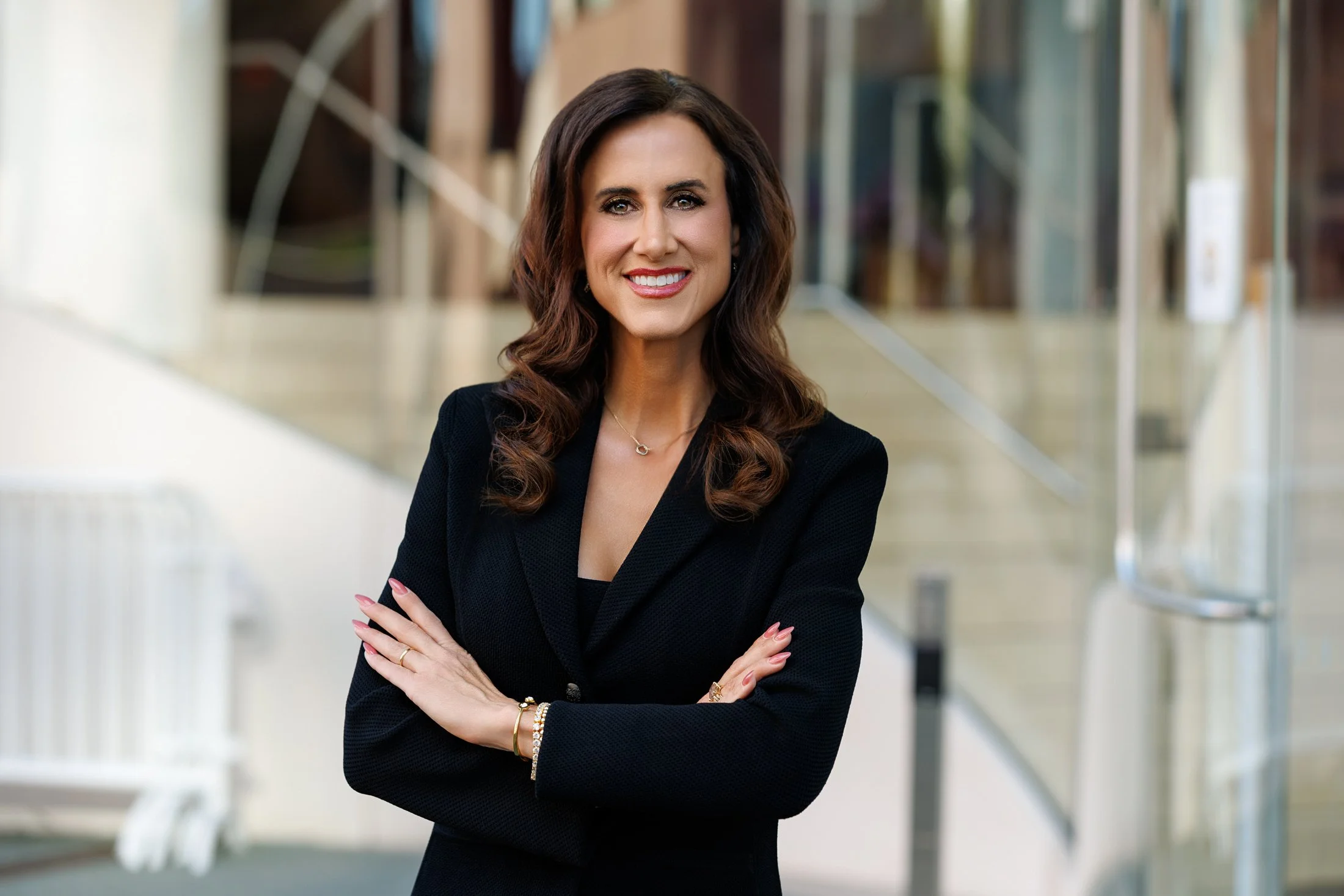 A woman with long auburn hair, wearing a black top and gold hoop earrings, standing outdoors with blurred green foliage in the background.