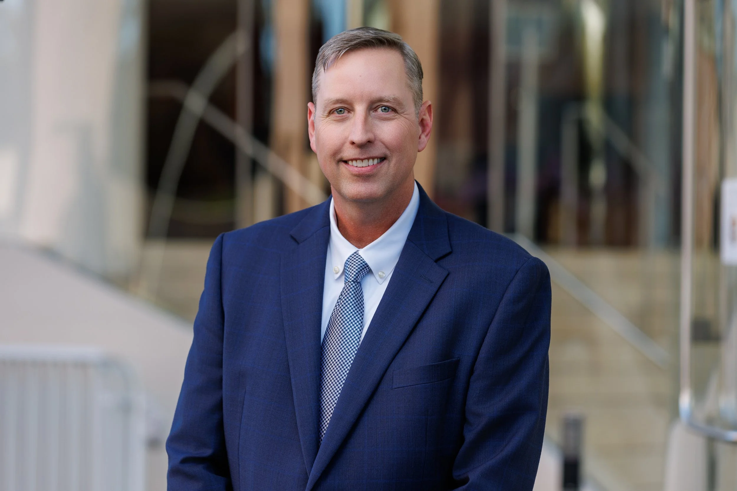 Headshot of a smiling man wearing a white dress shirt and a blue plaid tie, outdoors with blurred trees in the background.
