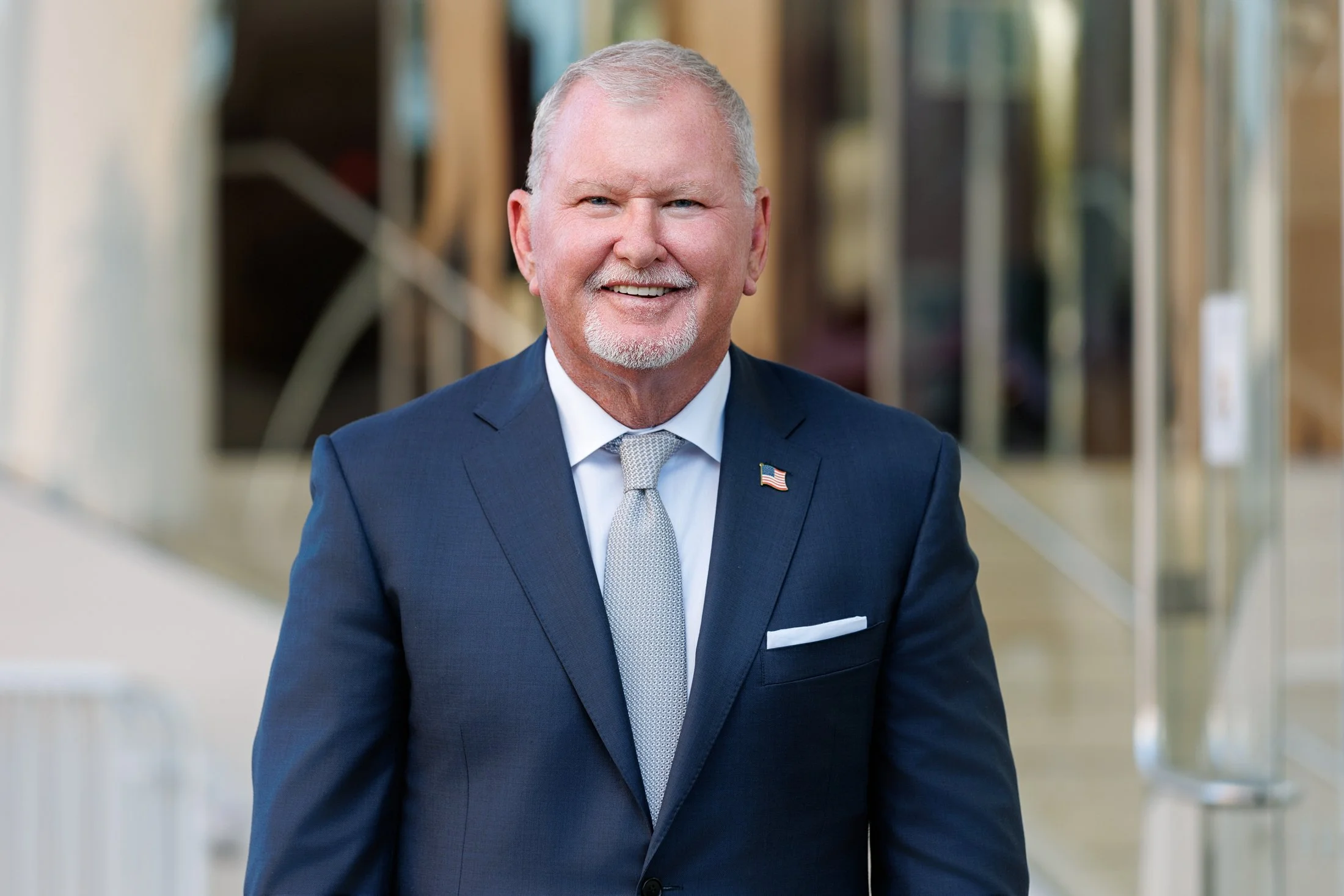 A smiling man with light skin, white hair, and a white beard, wearing a navy blue pinstripe suit, white dress shirt, and a small American flag lapel pin. He is outdoors with blurred background of buildings and trees.