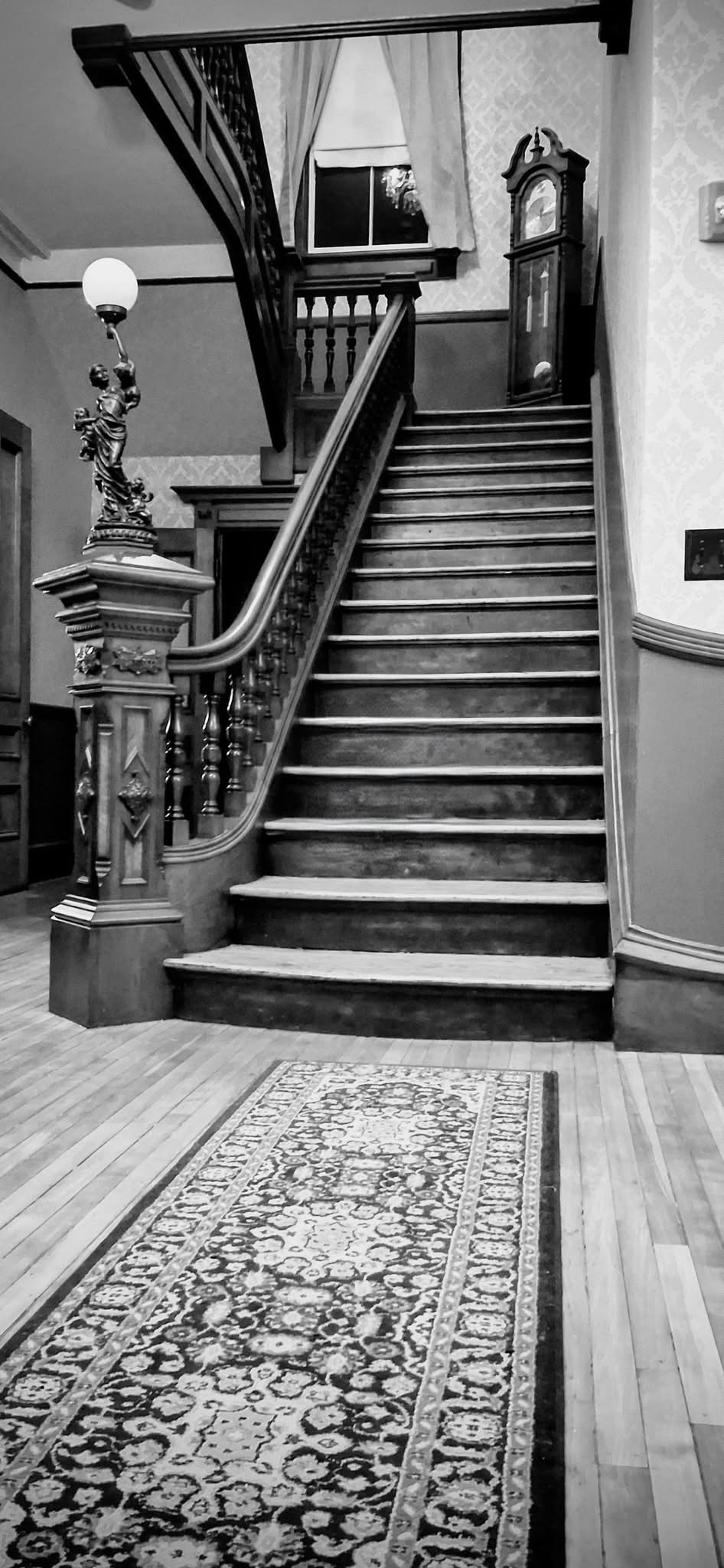 A black and white photo of a vintage staircase in the S.K. Pierce Mansion with a carpet runner, ornate wooden railing, a grandfather clock at the top, a decorative lamp with a figure holding a lantern, and a window with curtains.