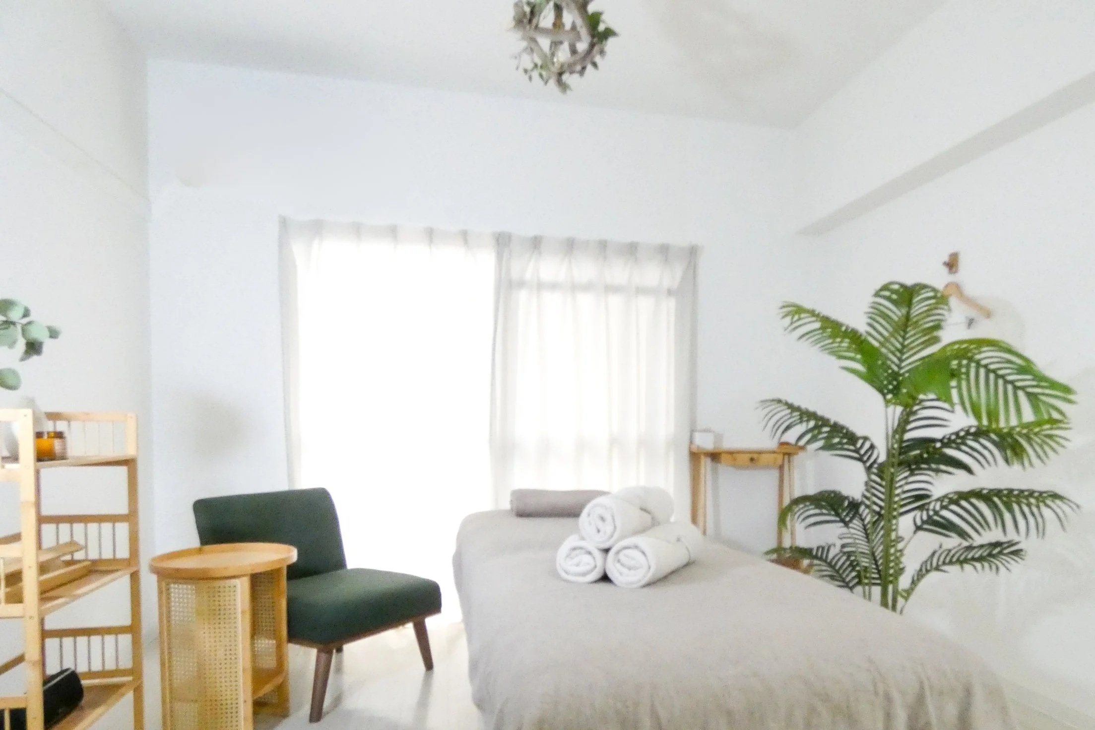 白い壁とカーテンのある明るい部屋。緑の観葉植物、木製の棚、椅子とベッドに白いタオルが巻かれている。