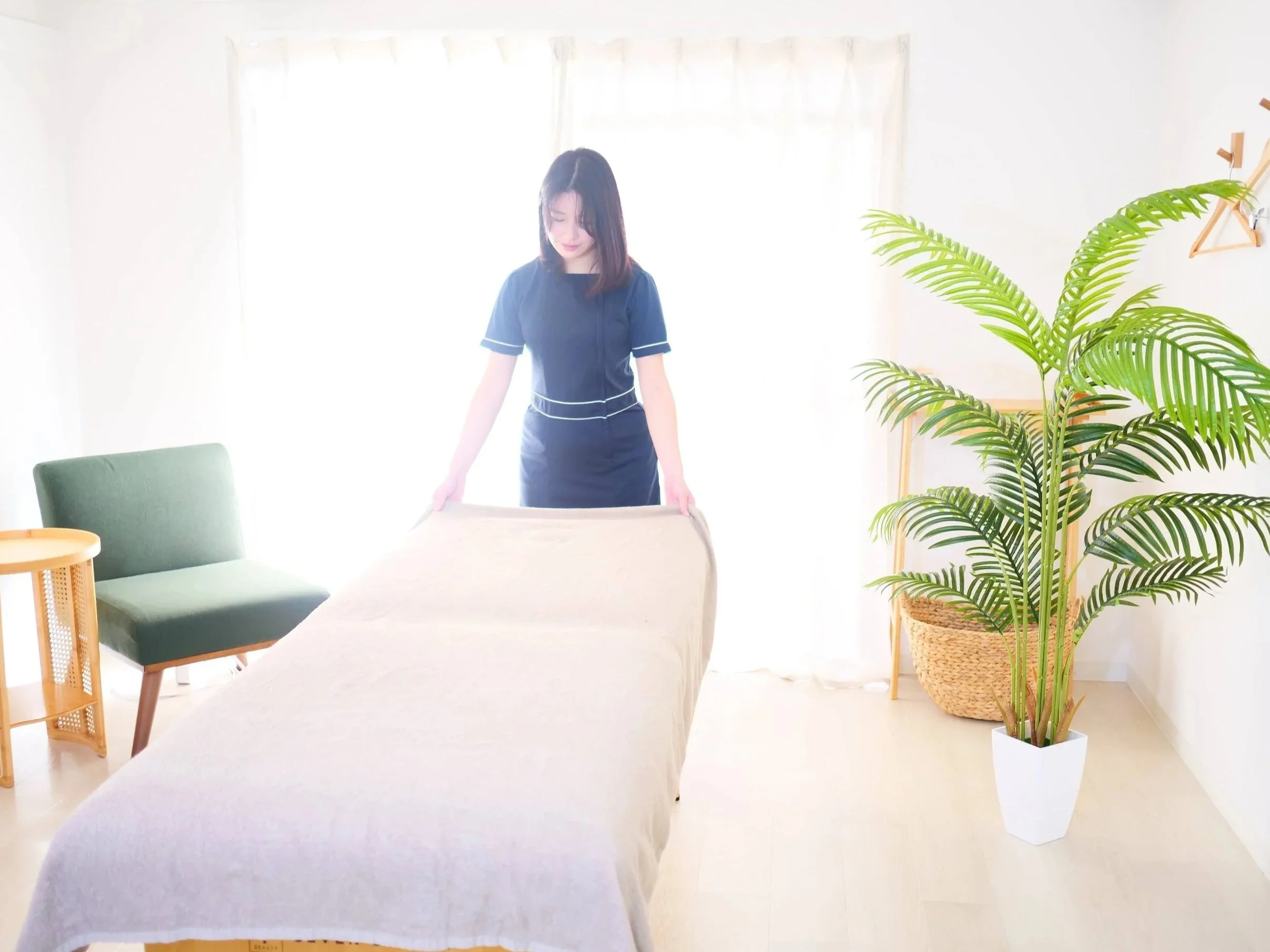 女性がベッドのシーツを整えている。室内には緑色の椅子と大きな観葉植物が置かれている。