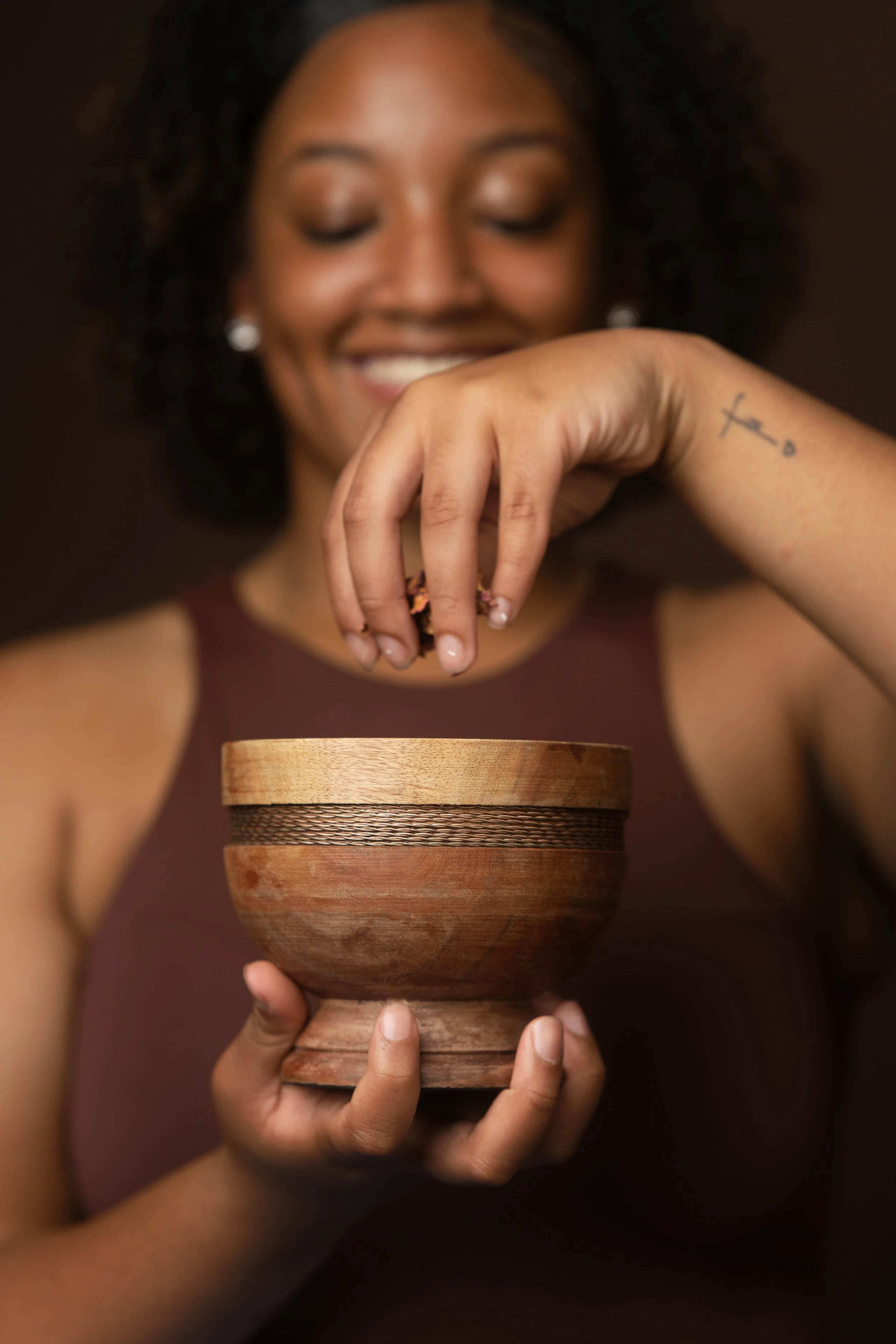 A smiling woman with curly hair holding a wooden bowl, sprinkling something into it with her right hand, and wearing a sleeveless brown top.
