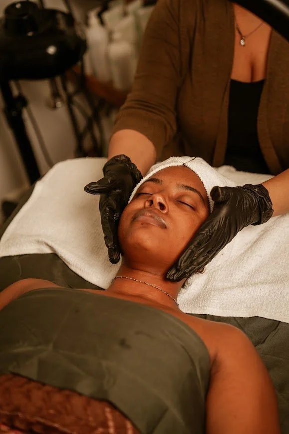 A woman receiving a facial treatment at a spa or skincare clinic, lying on a bed with her eyes closed, while a skincare professional wearing black gloves gently massages her face.