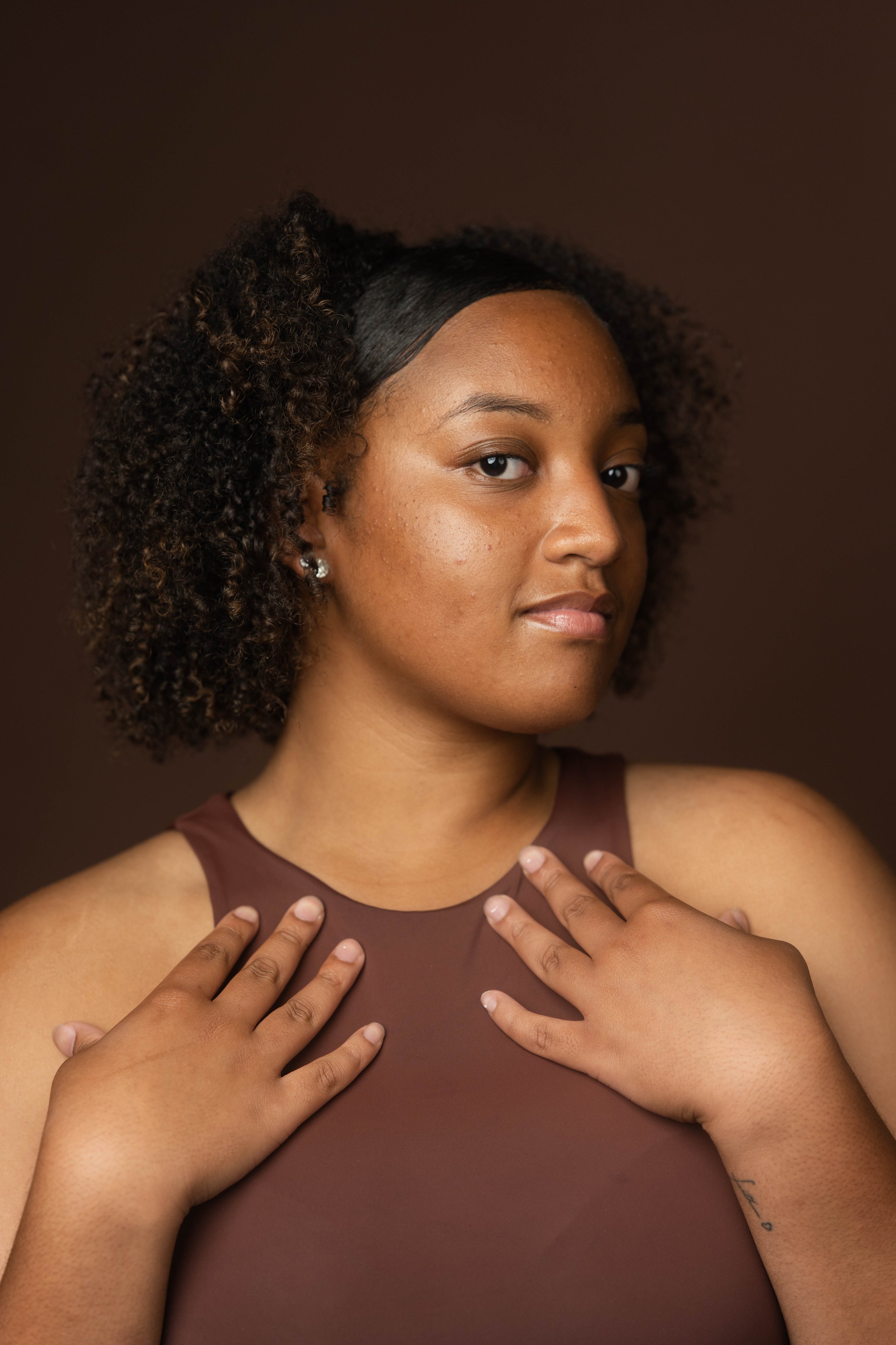 A woman with curly hair, wearing a brown sleeveless top, looking confidently at the camera with her hands placed on her chest.