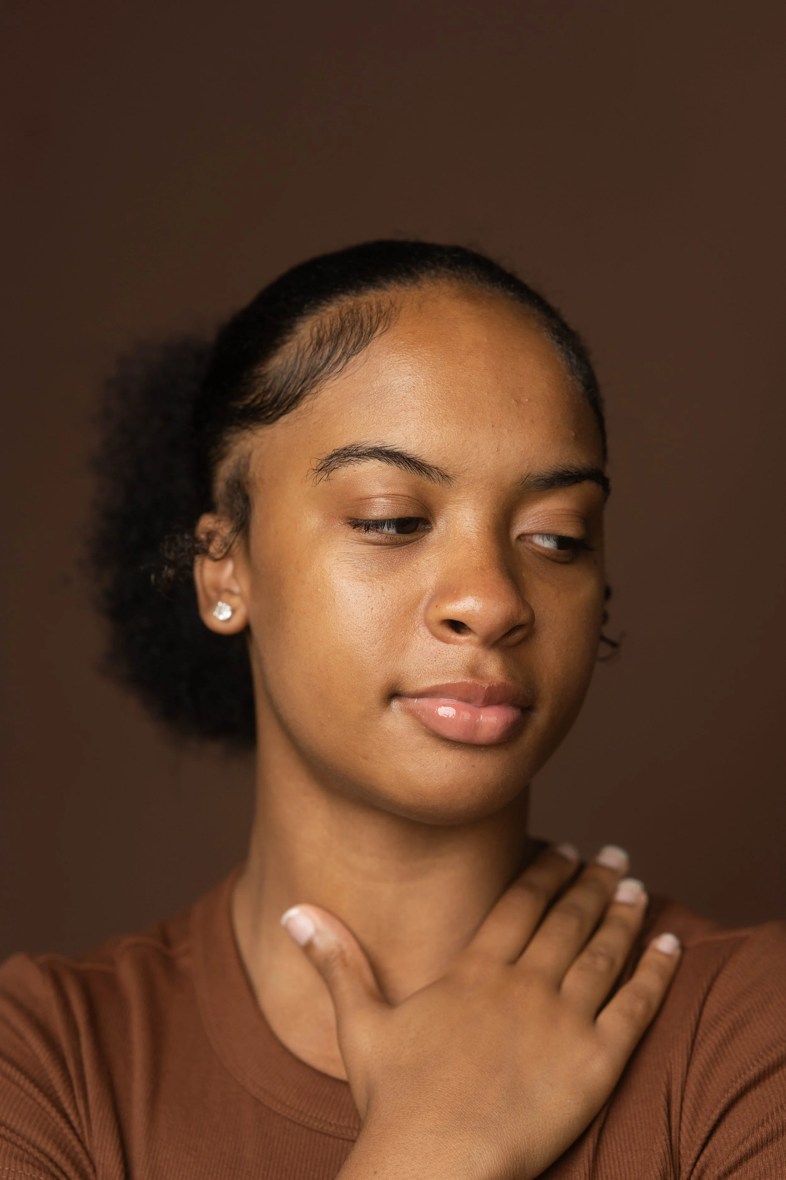 Close-up portrait of a young woman with natural hair, wearing earrings and a brown shirt, looking down with a gentle expression, against a brown background.