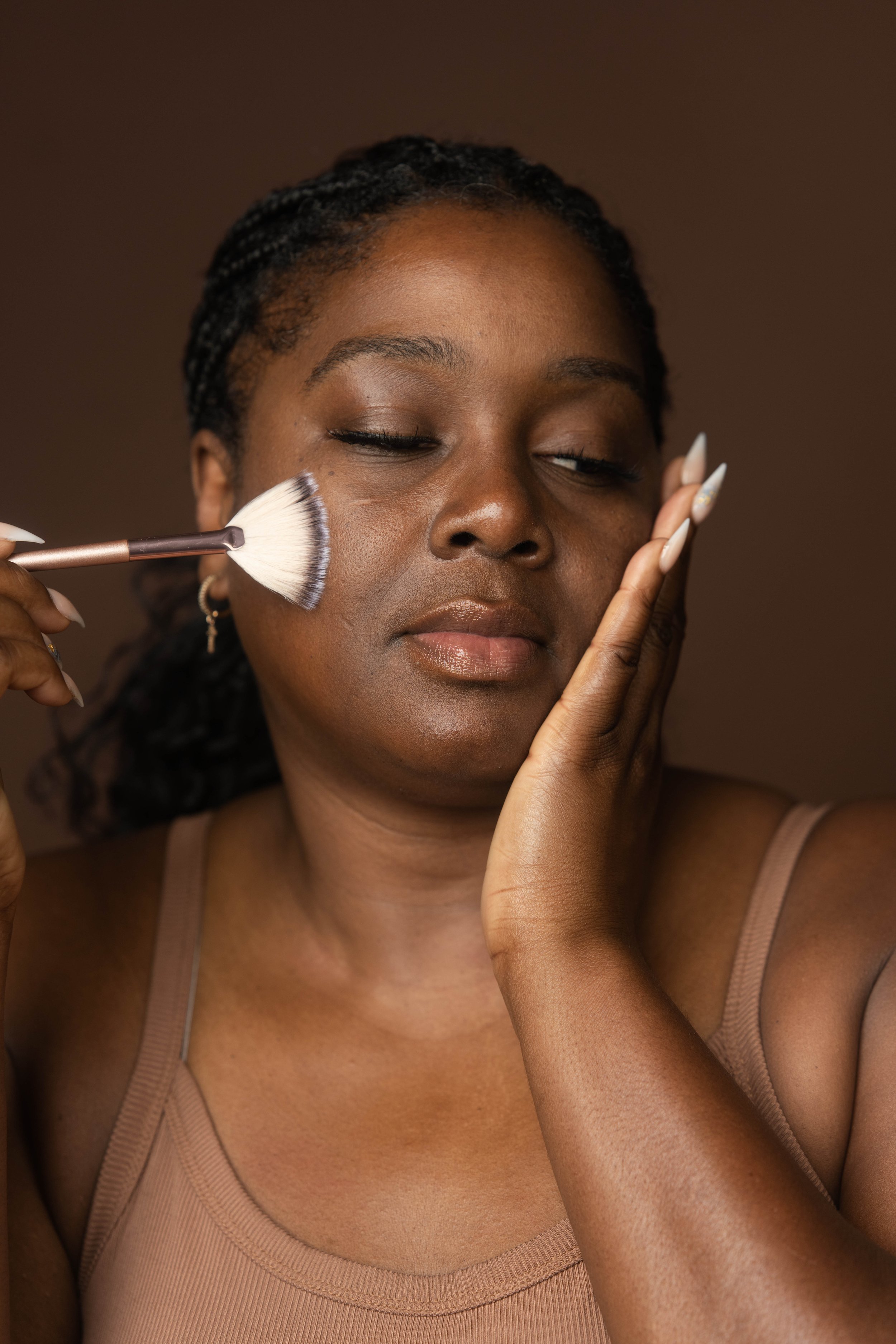 A woman with braided hair applies makeup to her face with a makeup brush, with her eyes closed and her hand resting on her cheek.