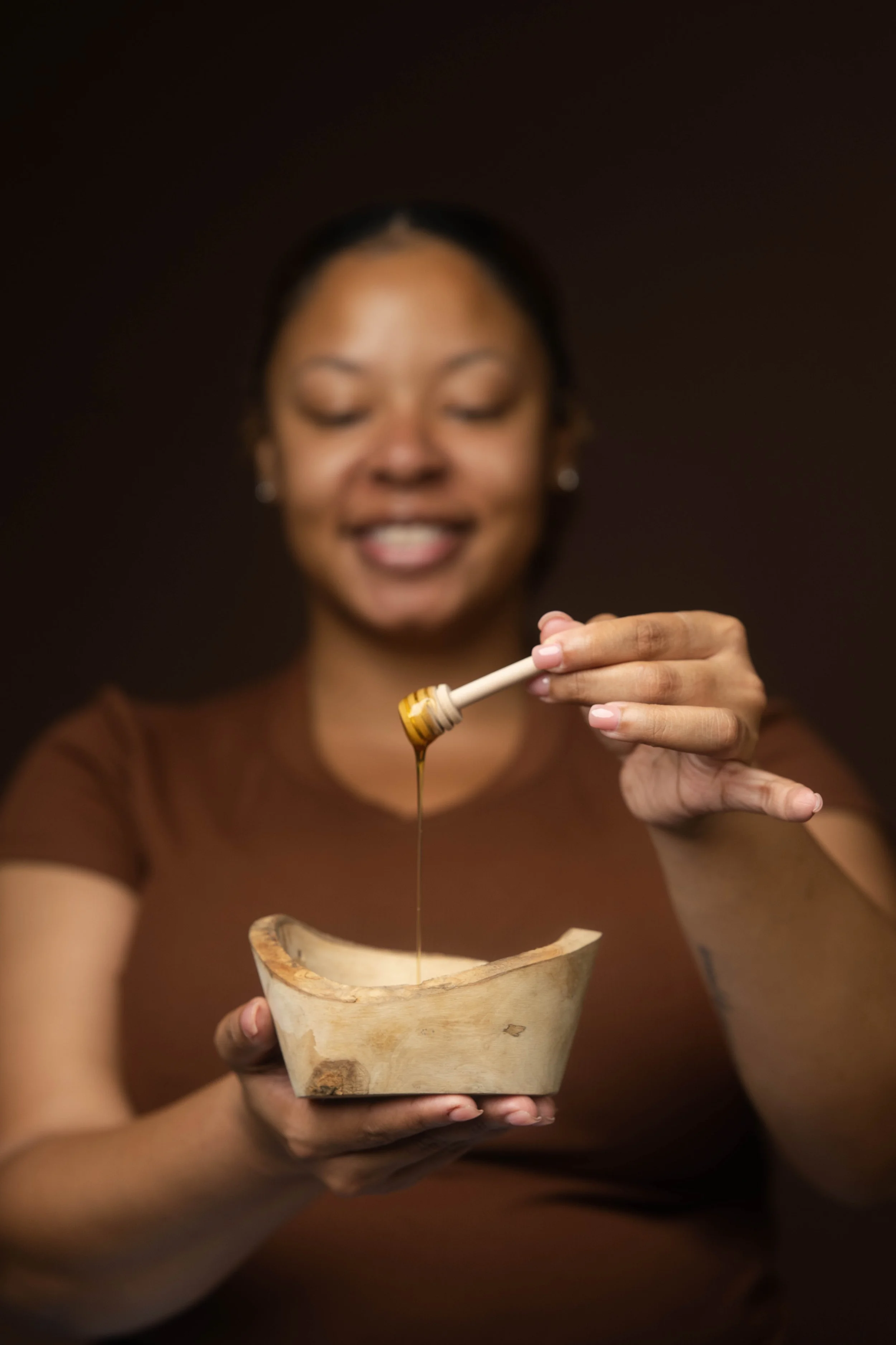 A woman is smiling and holding a small bowl while applying honey with a honey dipper.