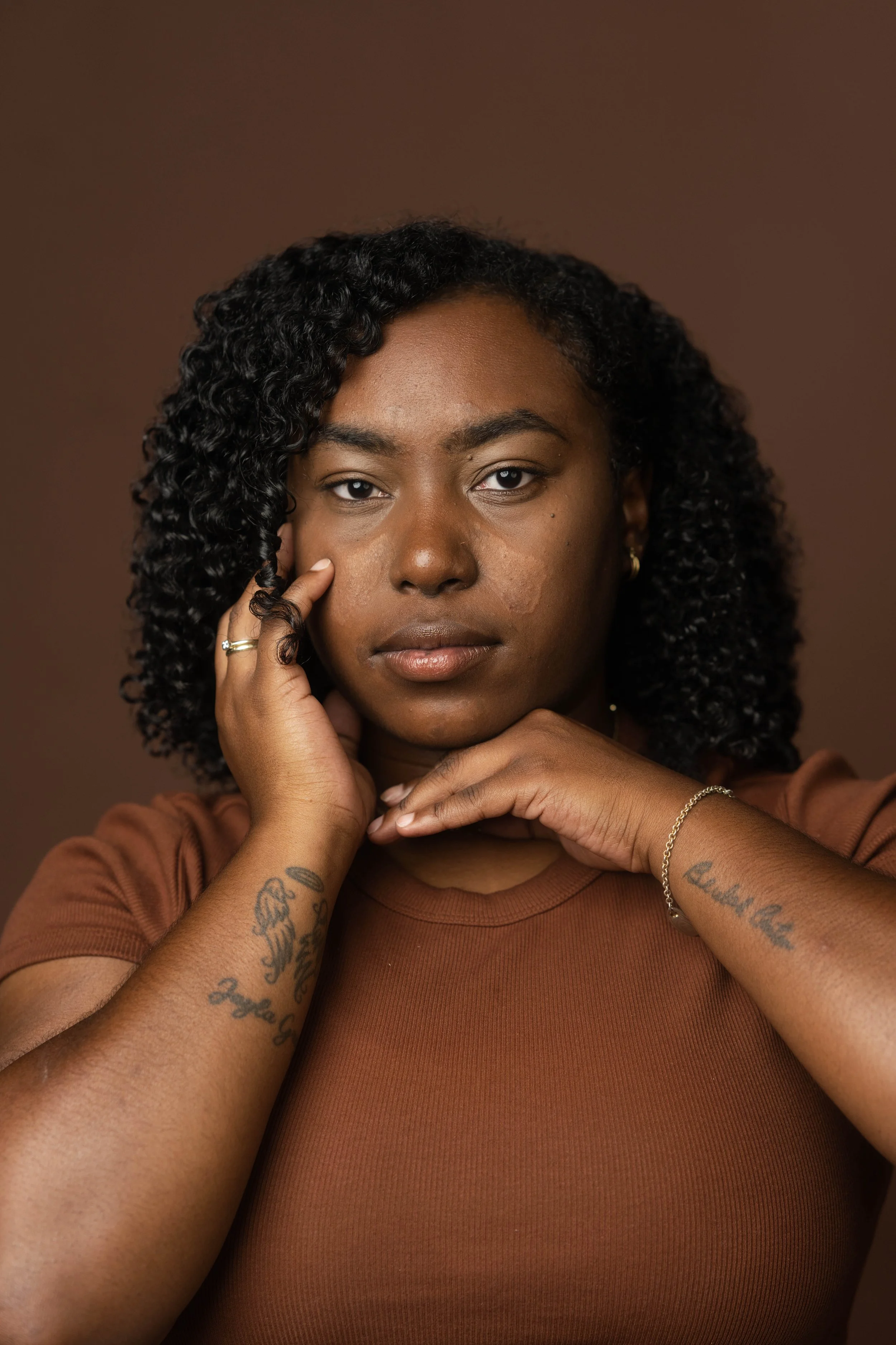 A woman with curly black hair and tattoos on her arms, wearing a brown shirt, looking directly at the camera with a serious expression, posing against a brown background.