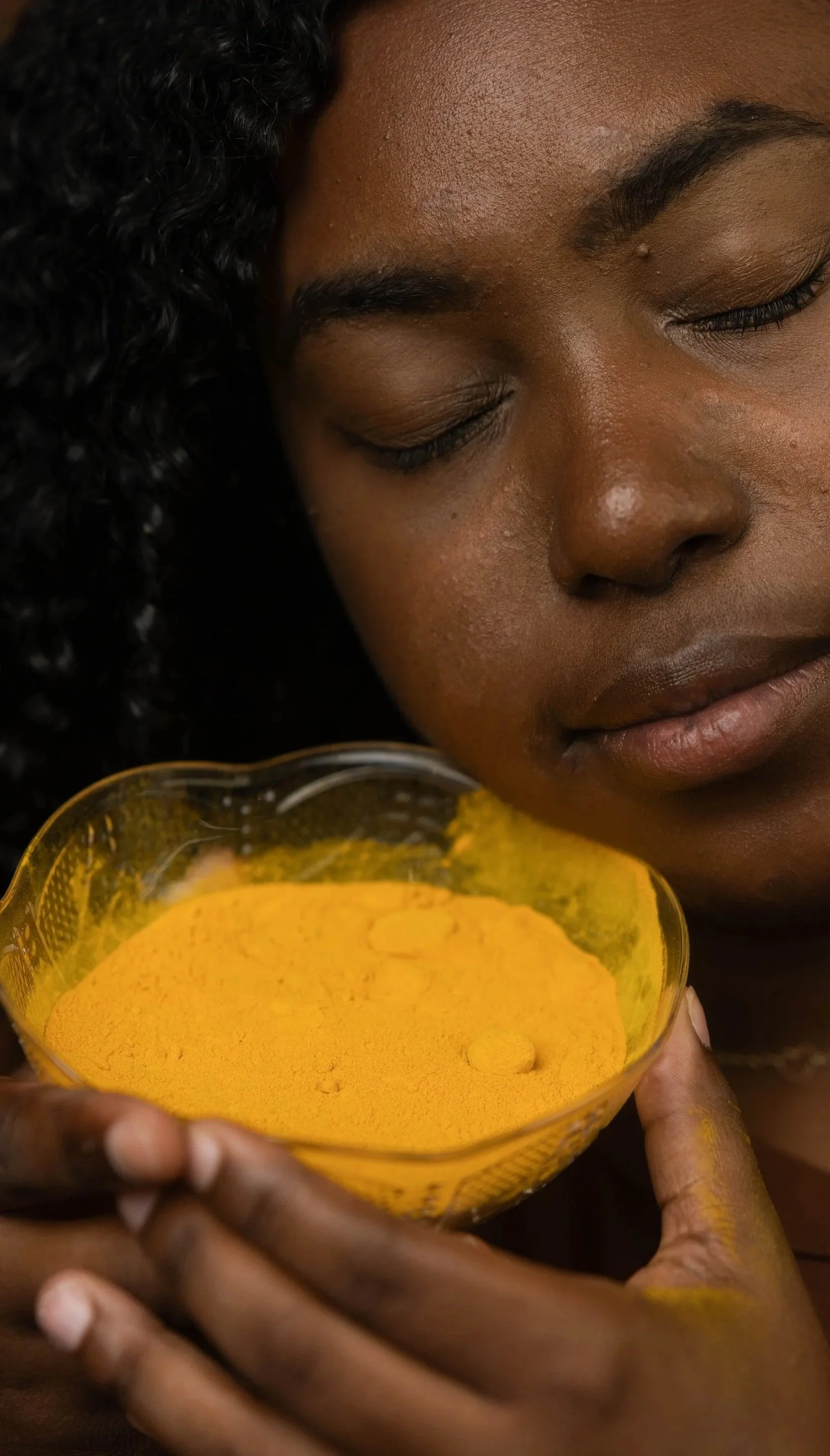 A woman with dark curly hair and dark skin holding a glass bowl filled with bright yellow powder, possibly turmeric, close to her face with eyes closed.