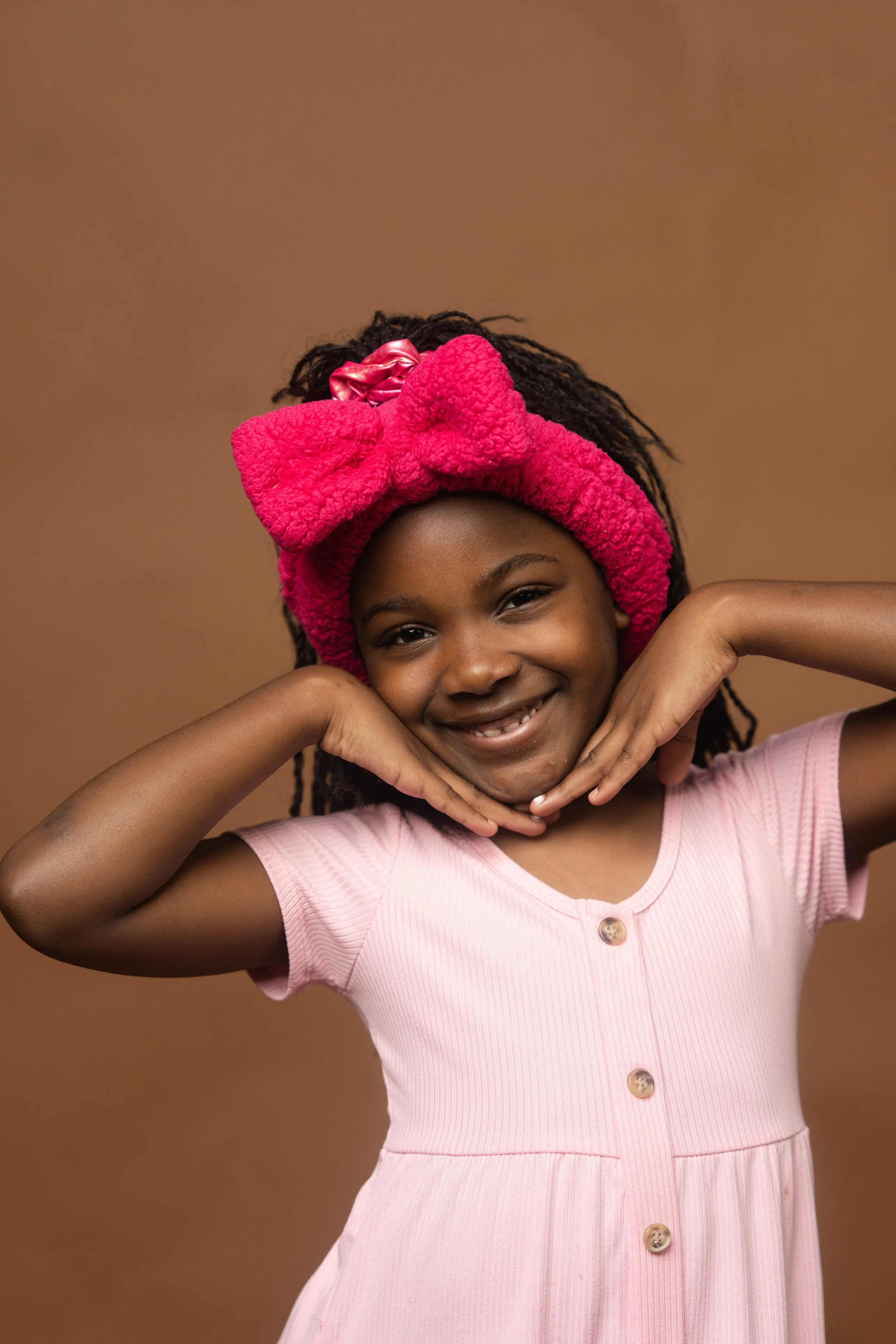 A young girl with a pink headband and pink dress, smiling and posing with her hands under her chin against a brown background.
