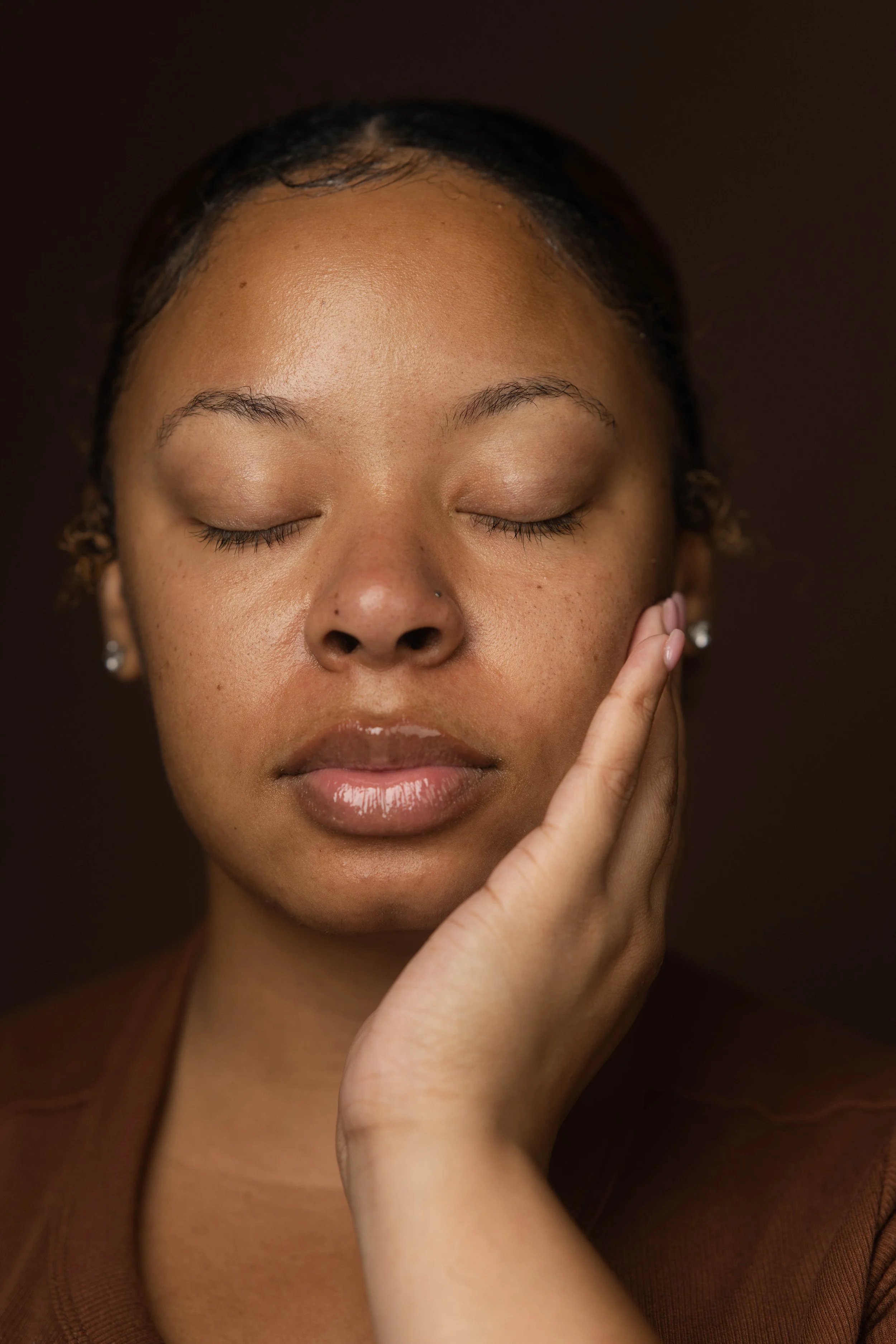 Close-up portrait of a woman with eyes closed, gently touching her face with one hand, black background.