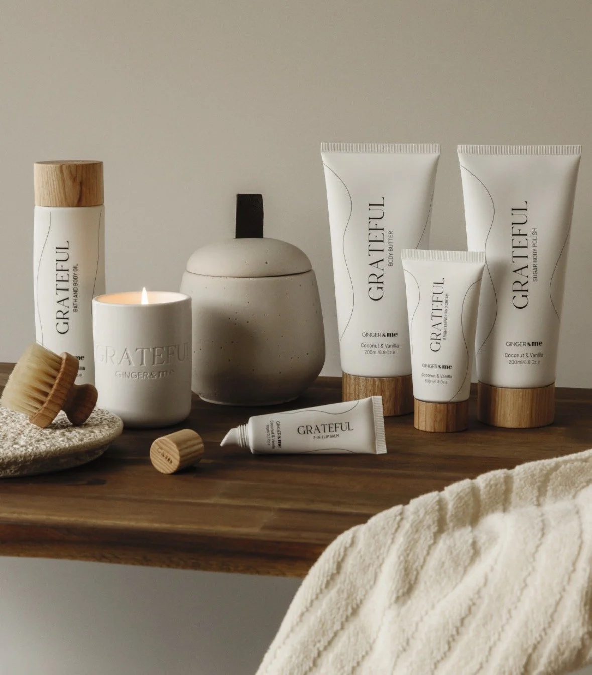 Collection of personal care products including body lotion, sugar body polish, hand cream, and lip balm labeled 'Grateful' on a wooden surface with a candle, wooden brushes, and a ceramic container.
