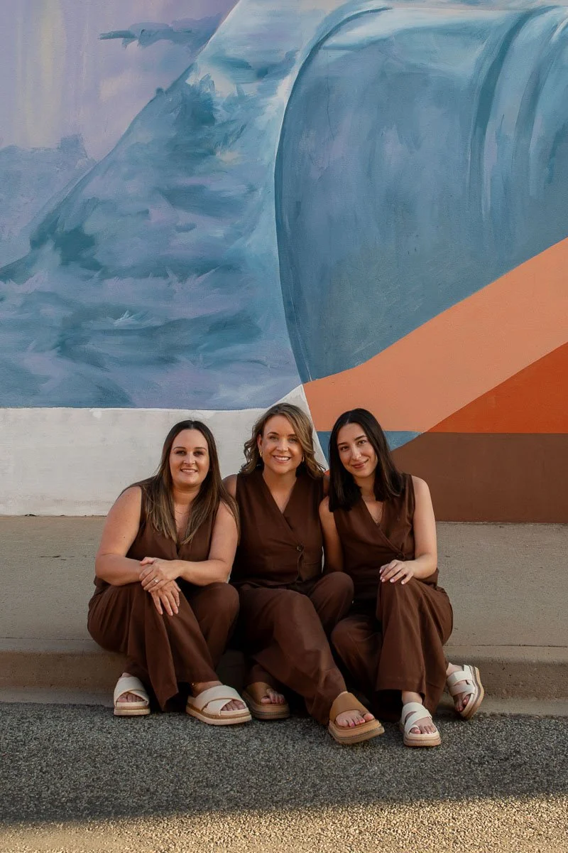 Three women sitting on the sidewalk in front of a colorful mural, smiling at the camera.