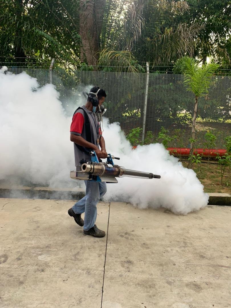 Man using a fogging machine to spray disinfectant outdoors, with smoke or fog emitting from the device, on concrete ground with trees and a fence in the background.