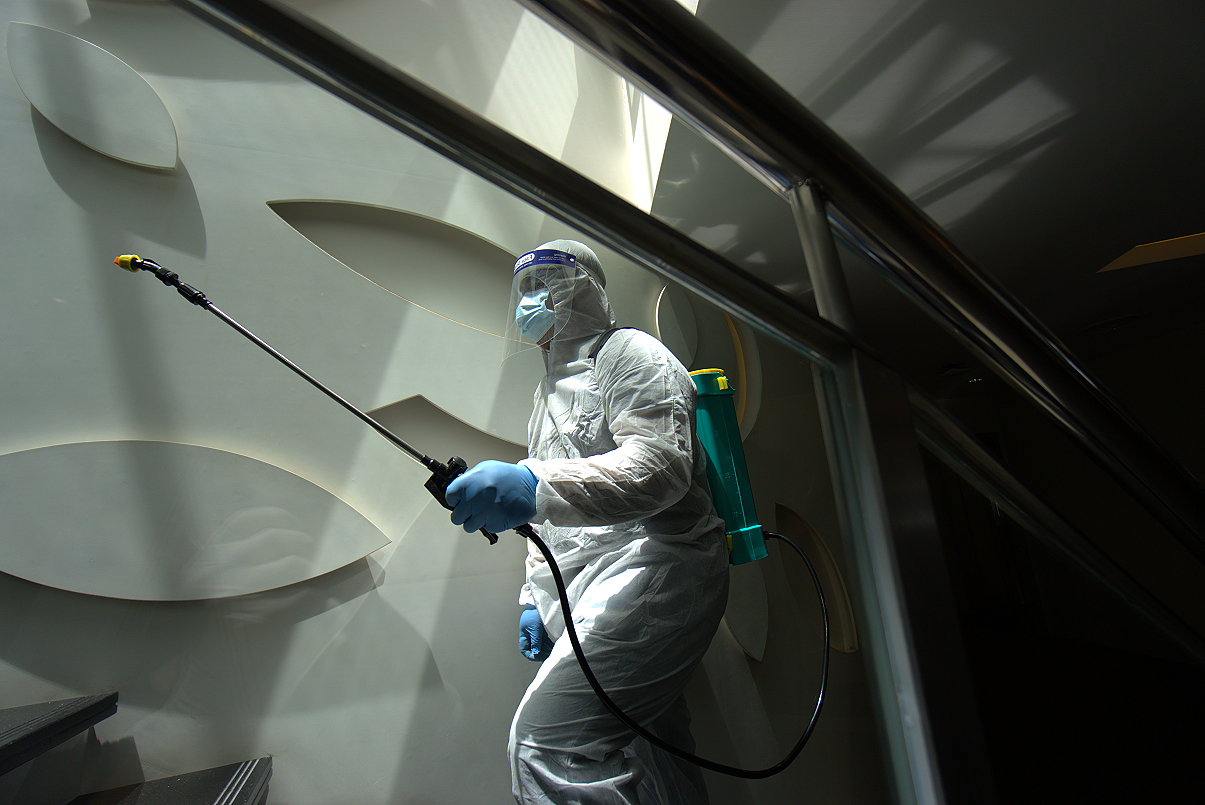 A person in full protective gear, including a face mask, face shield, gloves, and a hazmat suit, is disinfecting a wall with a sprayer.