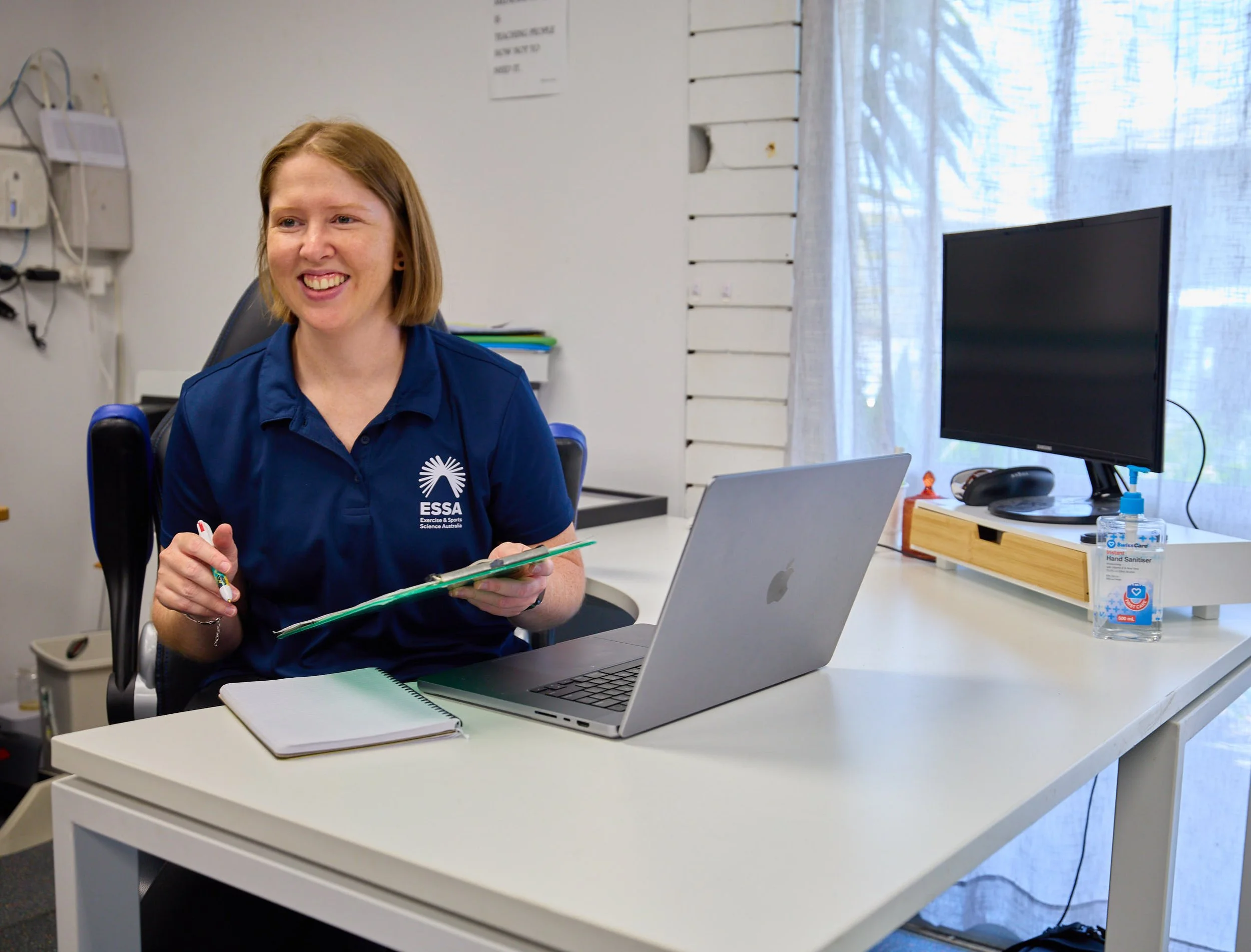 A woman in a blue shirt sitting at a white desk with a laptop, notebook, and clipboard, smiling at the camera, in an office with a large window and a bottle of hand sanitizer on the desk.