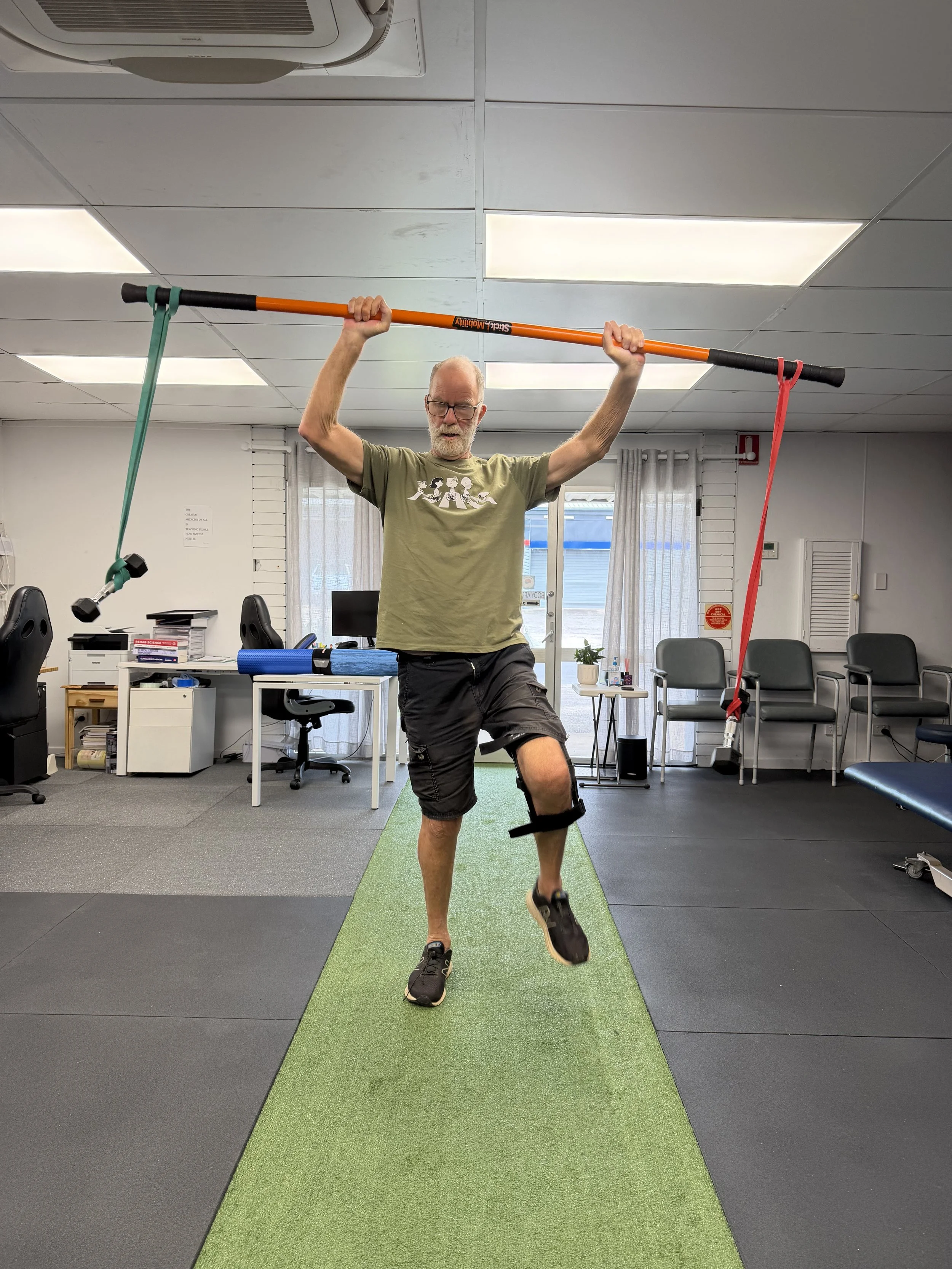 An elderly man with glasses performs a balance exercise in a therapy or rehabilitation room, holding a stick with resistance bands attached and balancing on one leg.