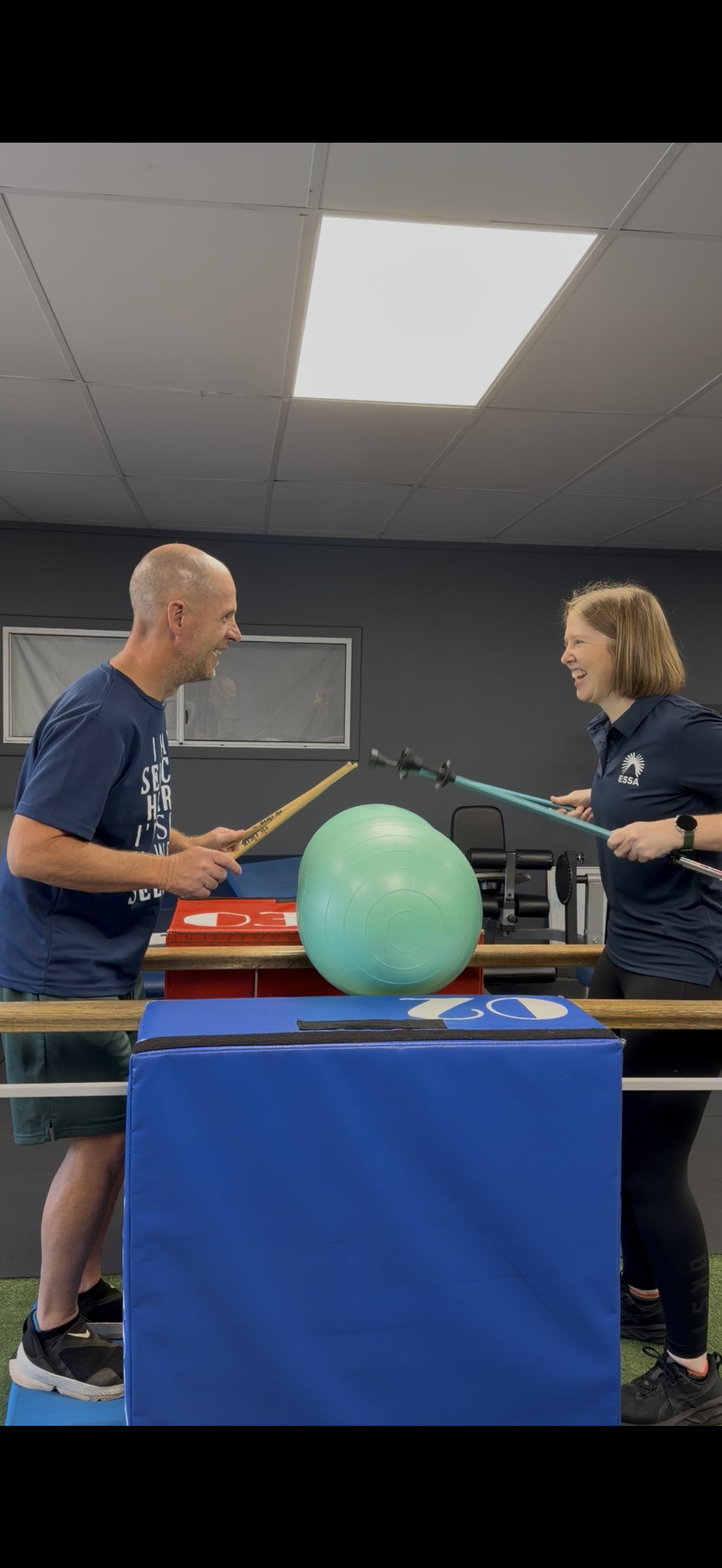 Two people standing behind a table with a green exercise ball, engaging in a playful fencing game with pool noodles. The man on the left is smiling, and the woman on the right is also smiling, both enjoying a fun activity.