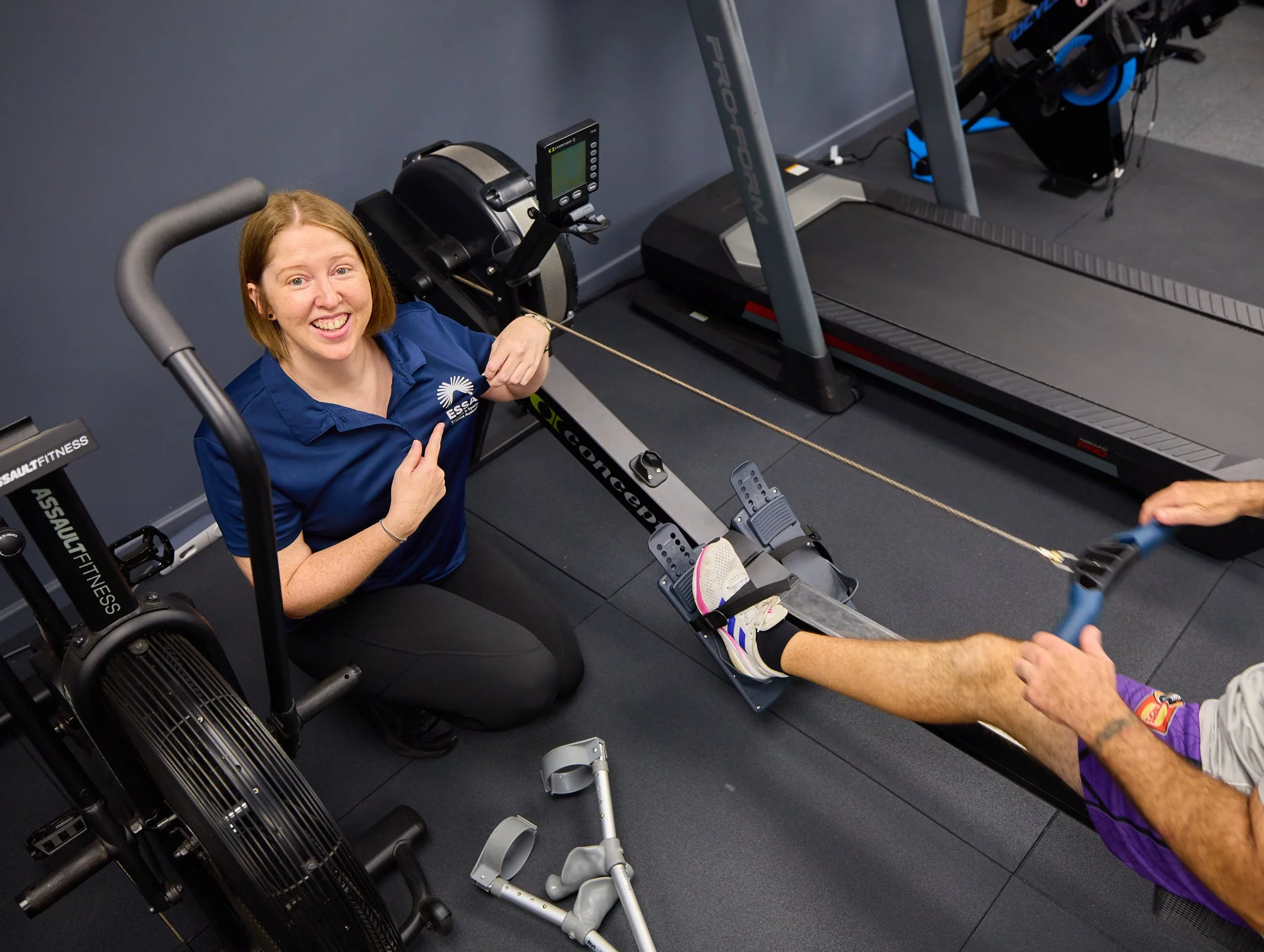 A woman in a blue shirt is posing and pointing at her jacket while seated on a rowing machine during a fitness assessment. A person in purple shorts is pulling on a resistance cord attached to her foot, which is secured in a footplate. The gym has treadmill and fitness equipment in the background.