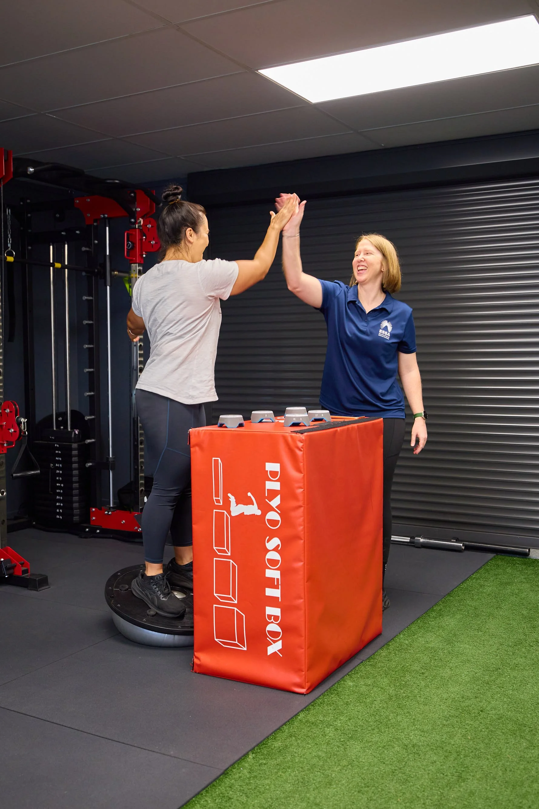 Two women high-fiving in a gym, one on a balance trainer and the other standing beside a red plyometric box labeled with blazepods on top.