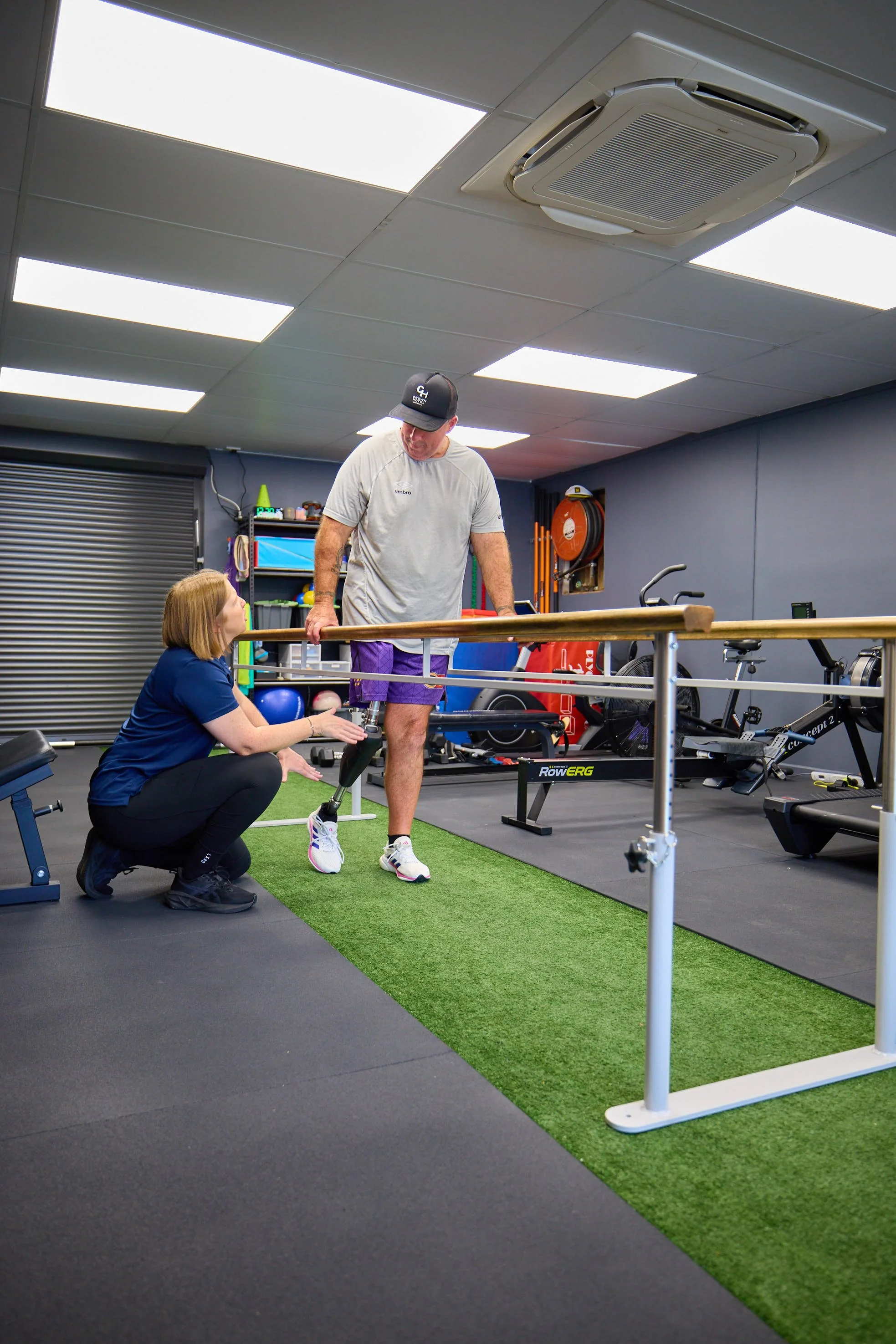 A man with a prosthetic leg doing physical therapy in a gym with a therapist assisting him, using parallel bars to practice walking.