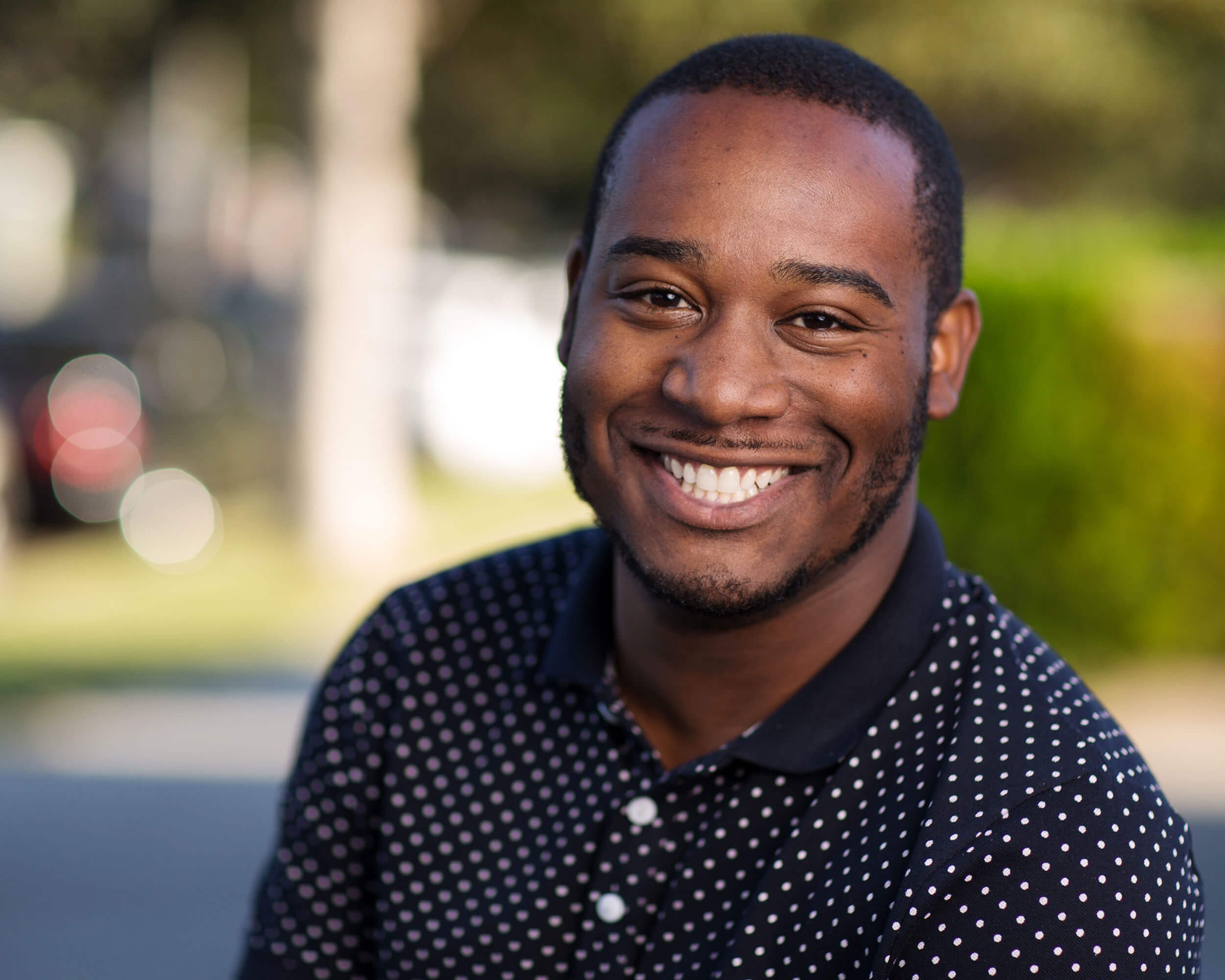 A smiling young man with short hair and a beard, wearing a black shirt with white polka dots, outdoors with a blurred background of trees and sunlight.