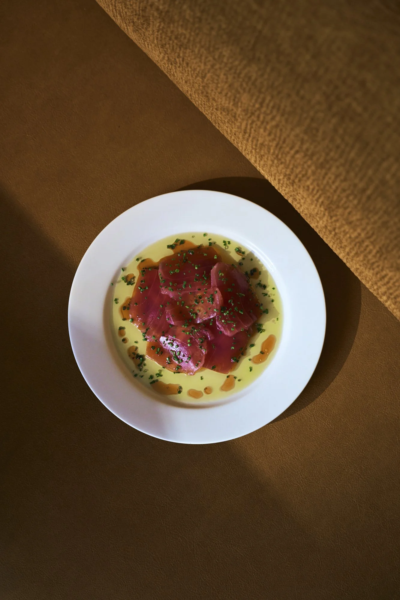 A white plate with slices of raw fish, likely tuna, garnished with chopped chives and drizzled with oil, on a brown surface.