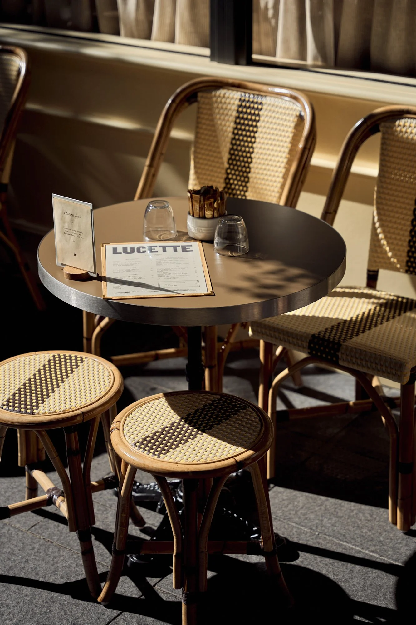 Outdoor café table with chairs, a menu, glasses, and a container of stir sticks, bathed in sunlight.