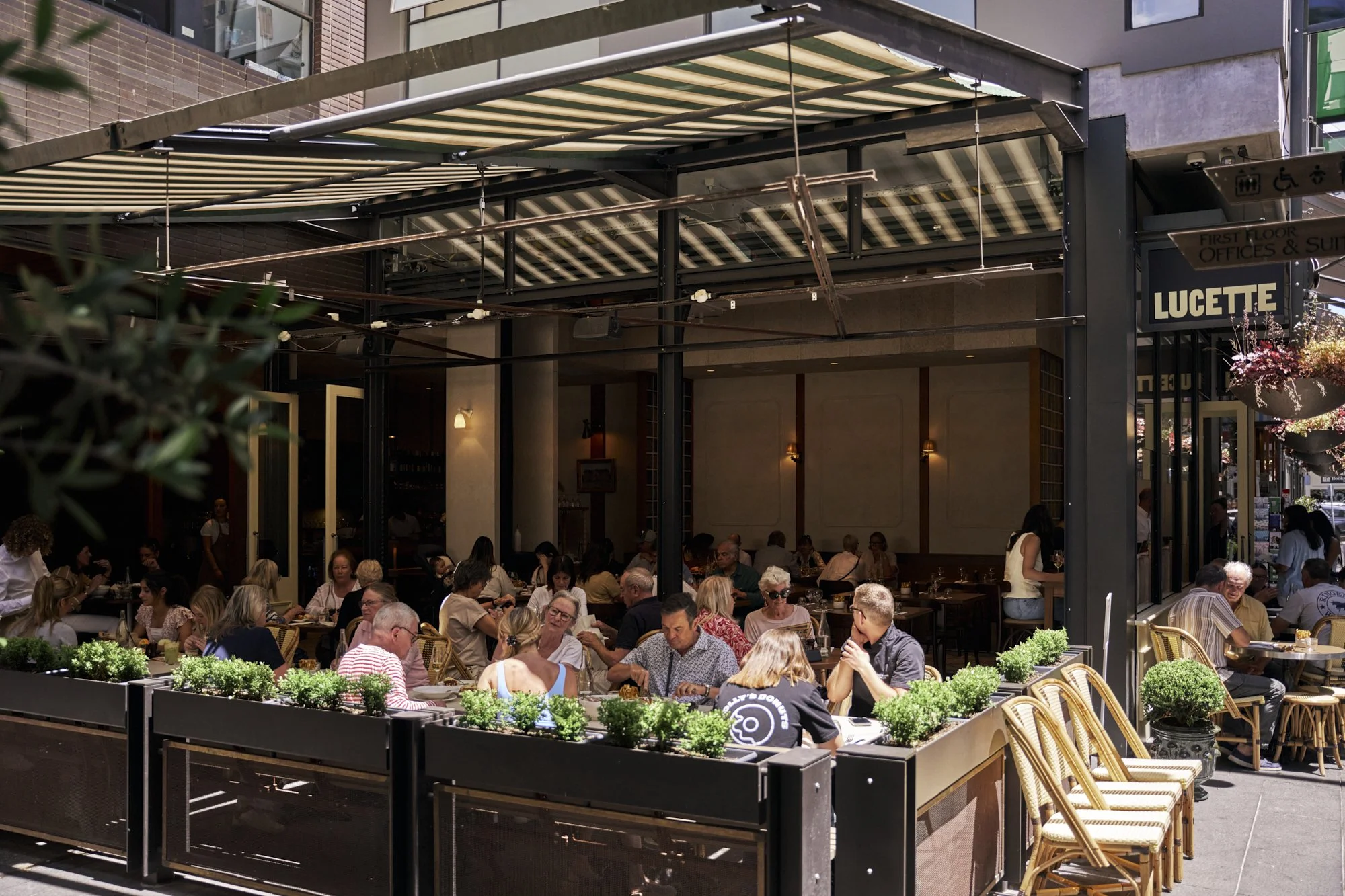 Outdoor seating area of a restaurant with many people dining, surrounded by potted plants and umbrellas, with some inside visible through open doors.