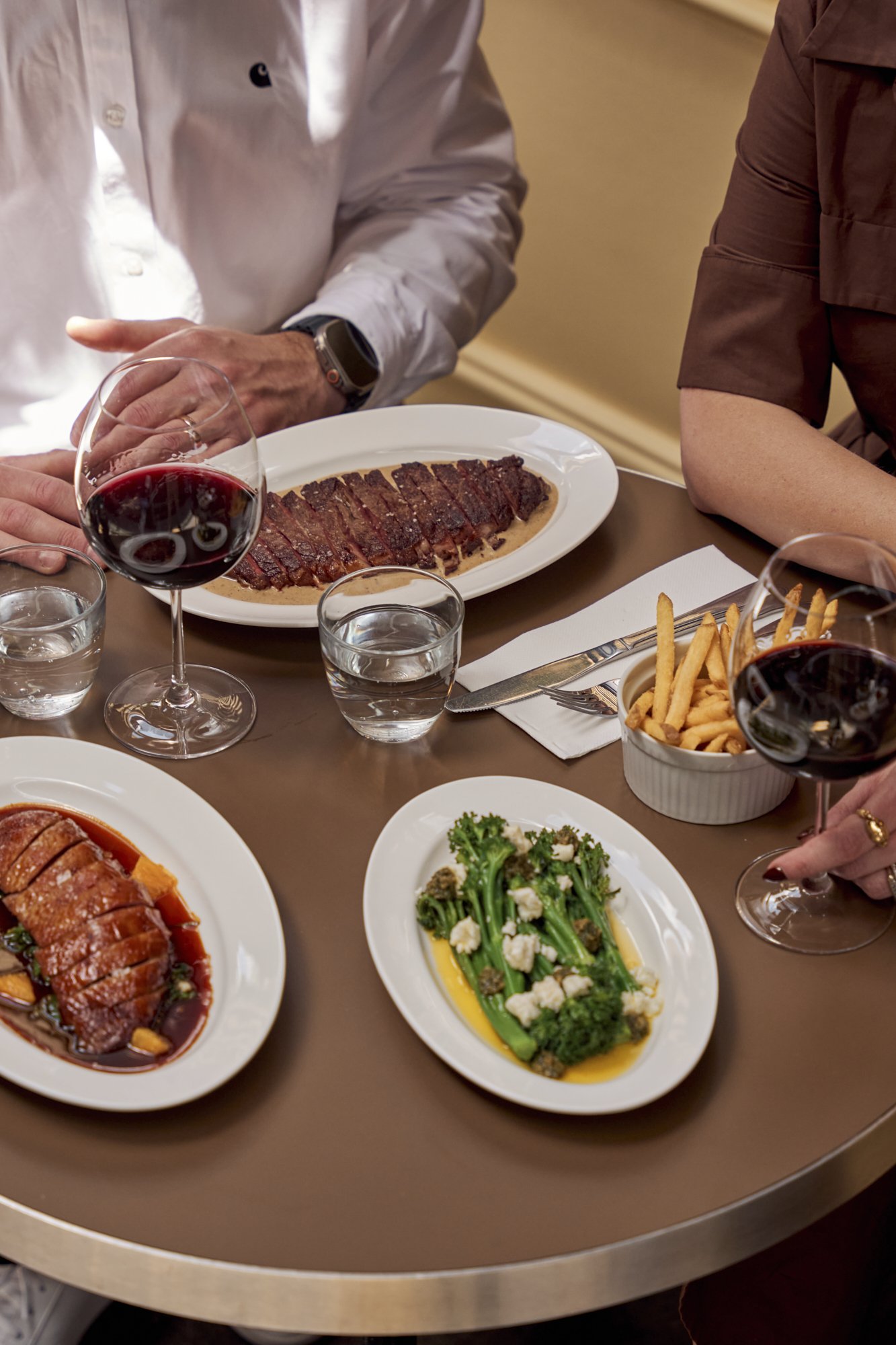 People dining at a restaurant with plates of steak, broccoli, fries, and glasses of red wine on the table.