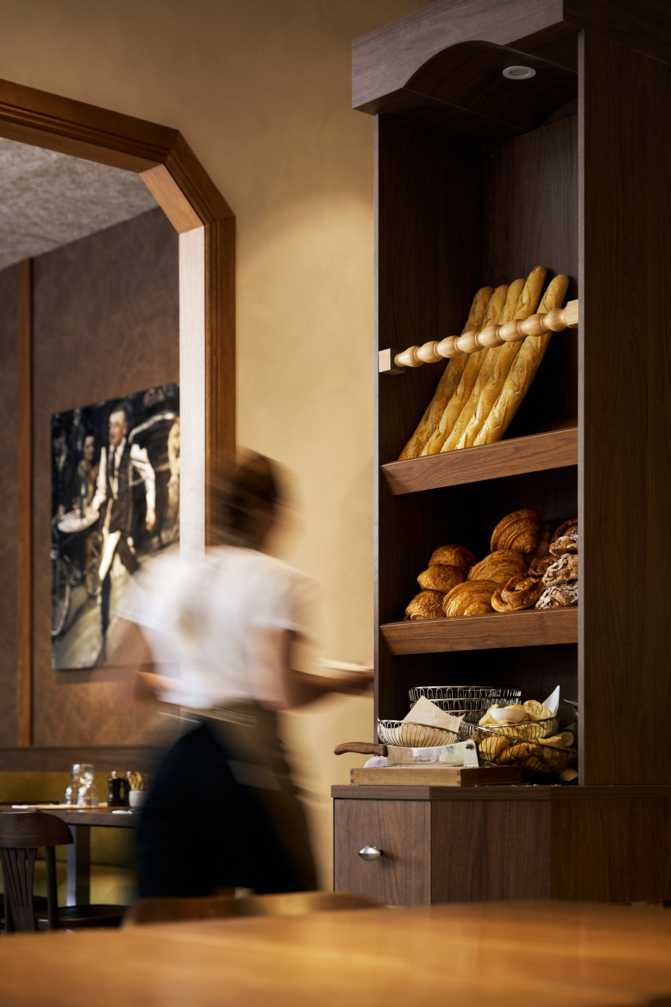 A wooden shelving unit with baguettes, croissants, and various bread rolls in a restaurant or cafe.