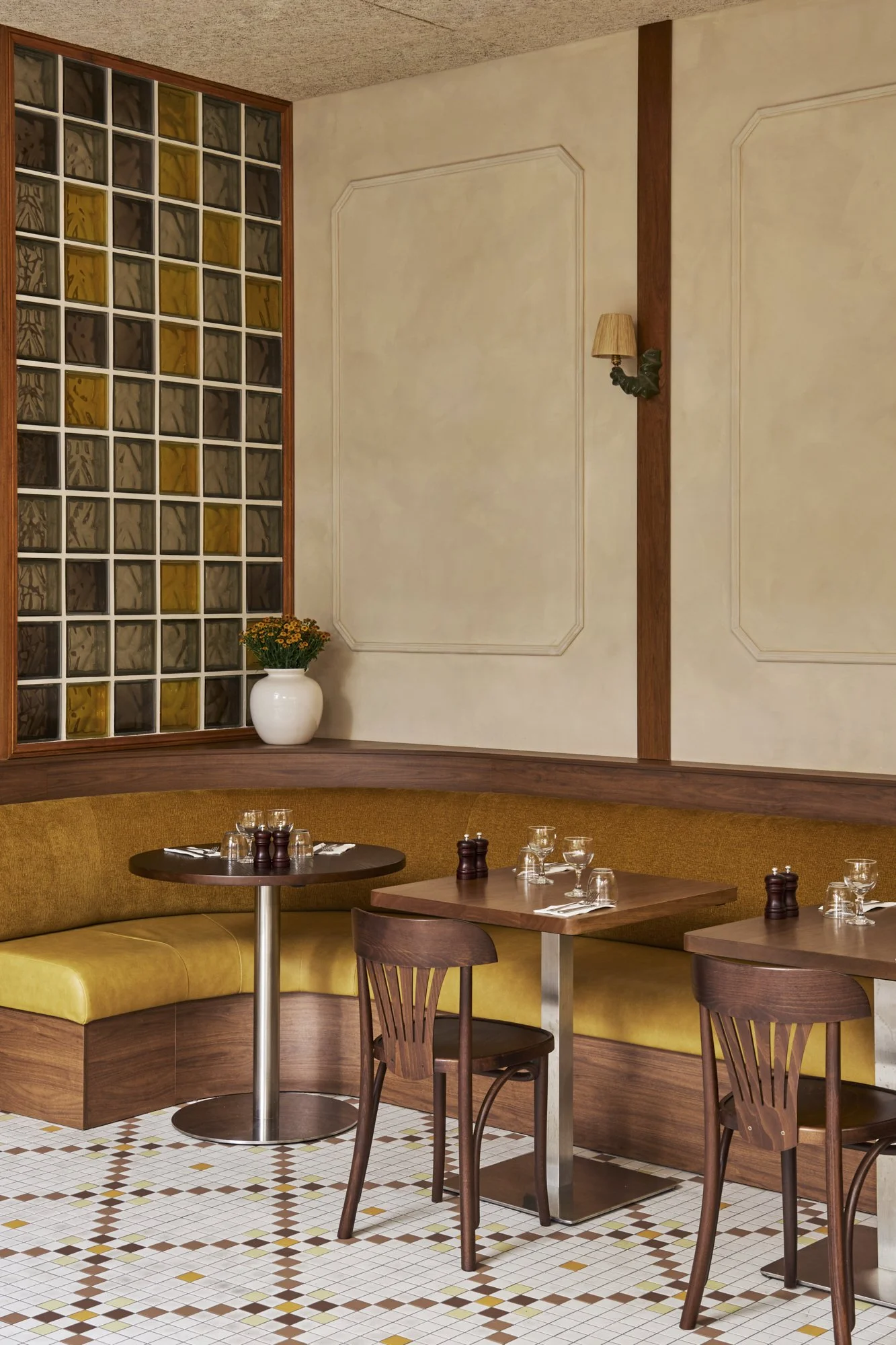 Interior of a restaurant with yellow cushioned booth seating, dark wooden tables, and chairs, white and brown tiled floor, a white vase with flowers on a ledge, and a decorative wall with a glass block window.