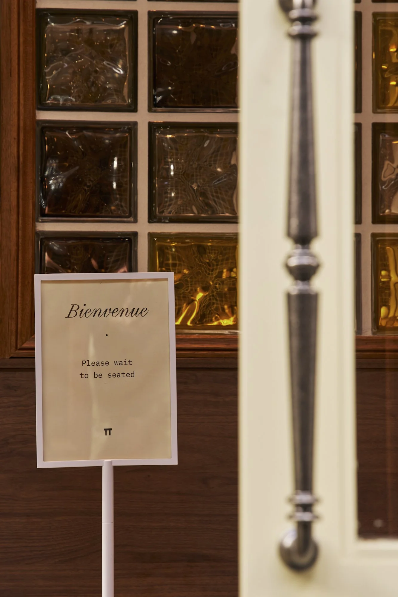 A sign in front of a wooden wall with glass block windows. The sign reads 'Bienvenue. Please wait to be seated' with a pi symbol at the bottom.