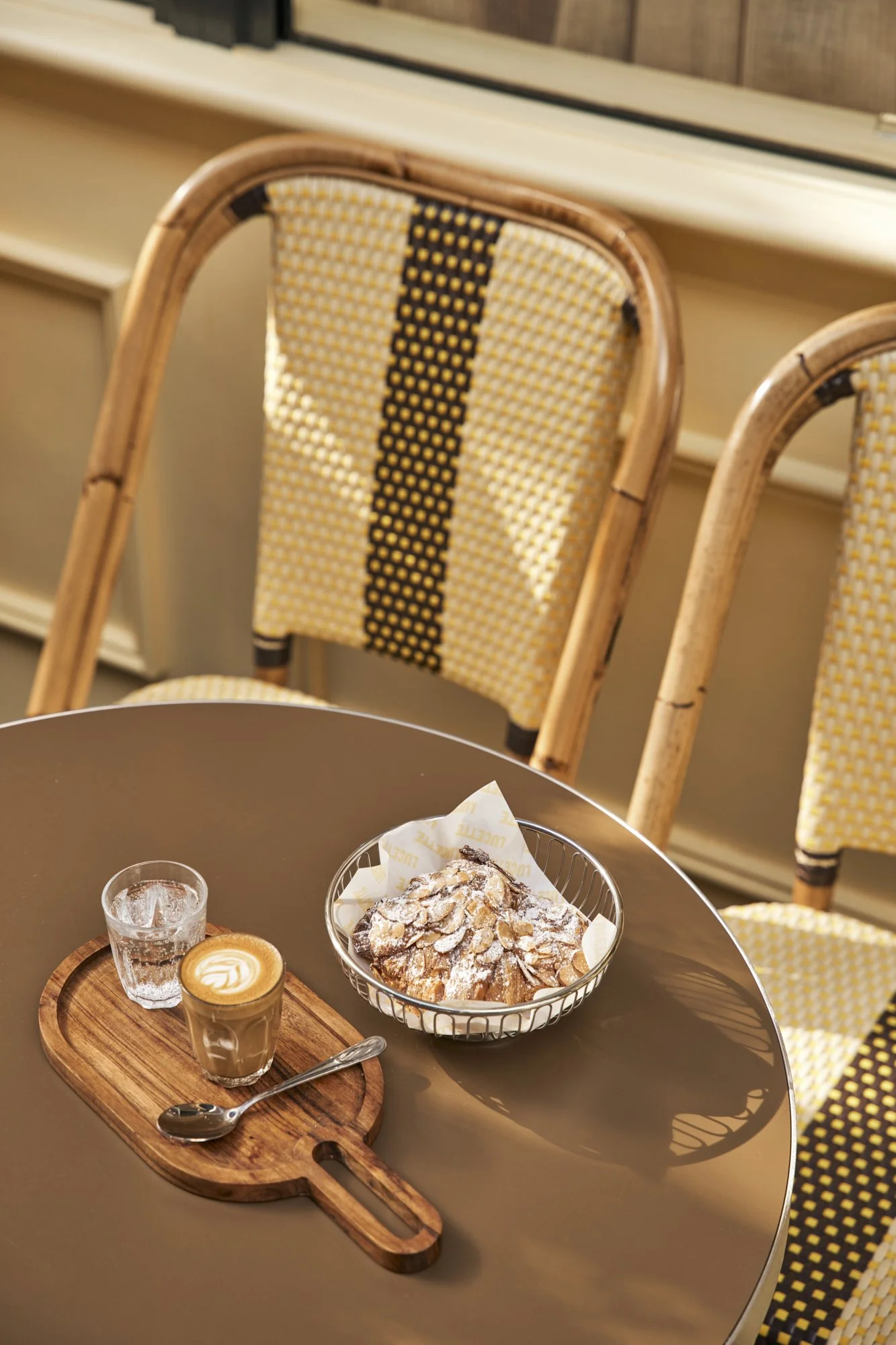 A round table with a wooden tray holding a glass of water, a glass of coffee with latte art, and a small spoon. Next to the tray is a basket of powdered sugar-dusted pastry topped with almonds. Two woven chairs with yellow and black patterned cushion