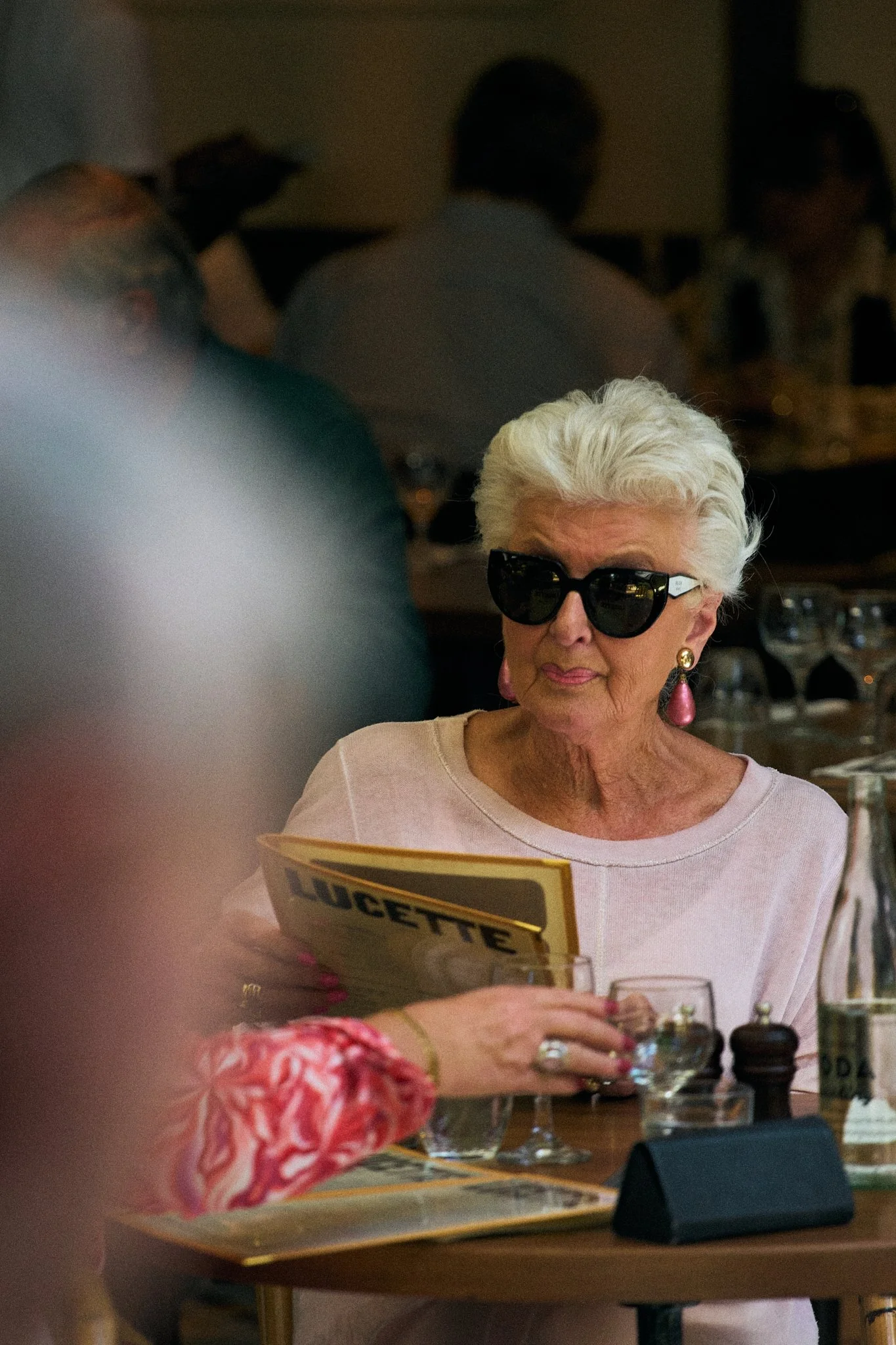 An elderly woman with white hair, wearing large black sunglasses and pink earrings, sitting at a table in a restaurant, looking at a menu.