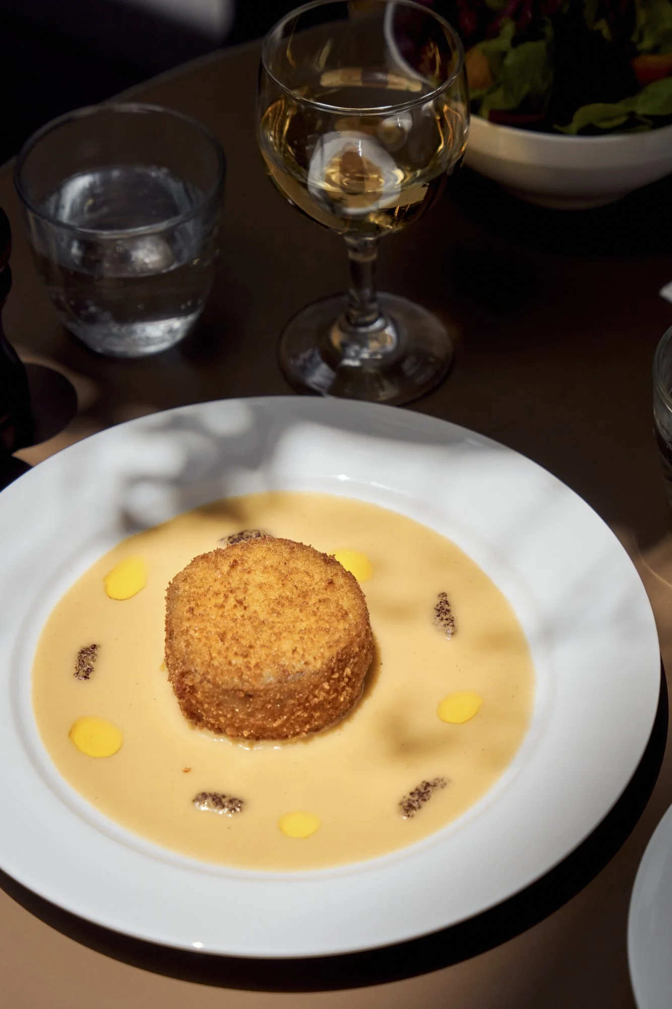 A plate with a breaded, fried appetizer in a light-colored sauce garnished with small yellow and dark crumbs, accompanied by a glass of white wine, a glass of water, and a bowl of salad in the background.