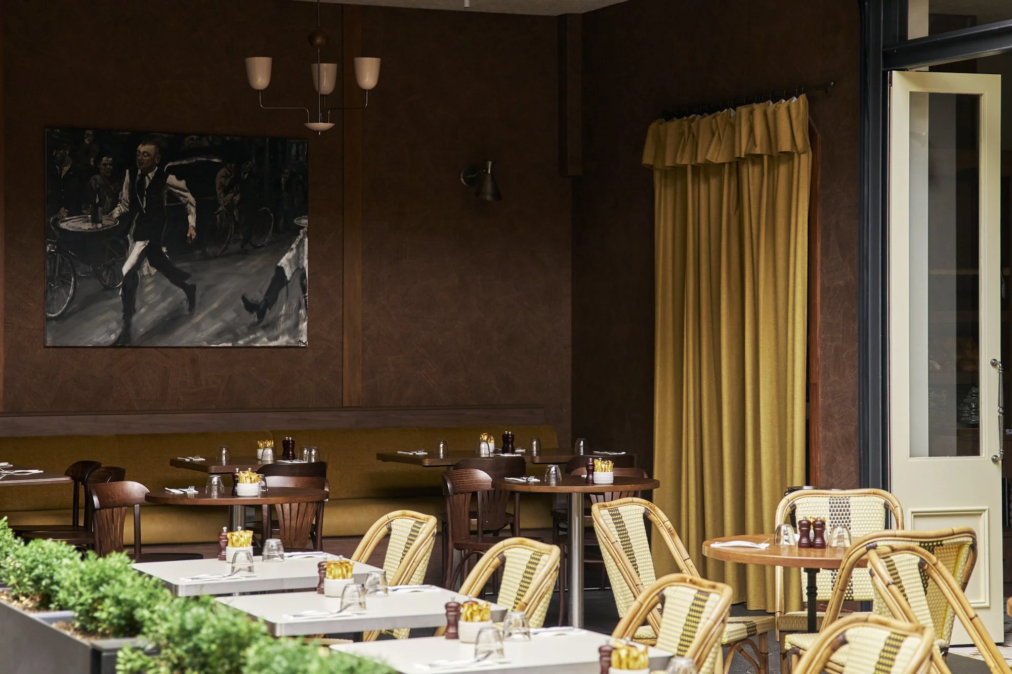 Interior of a restaurant with wooden and yellow seating, tables set with glasses, napkins, and condiments. A large painting of waiters and cyclists hangs on the wall, and a yellow curtain partially covers an open door or window.