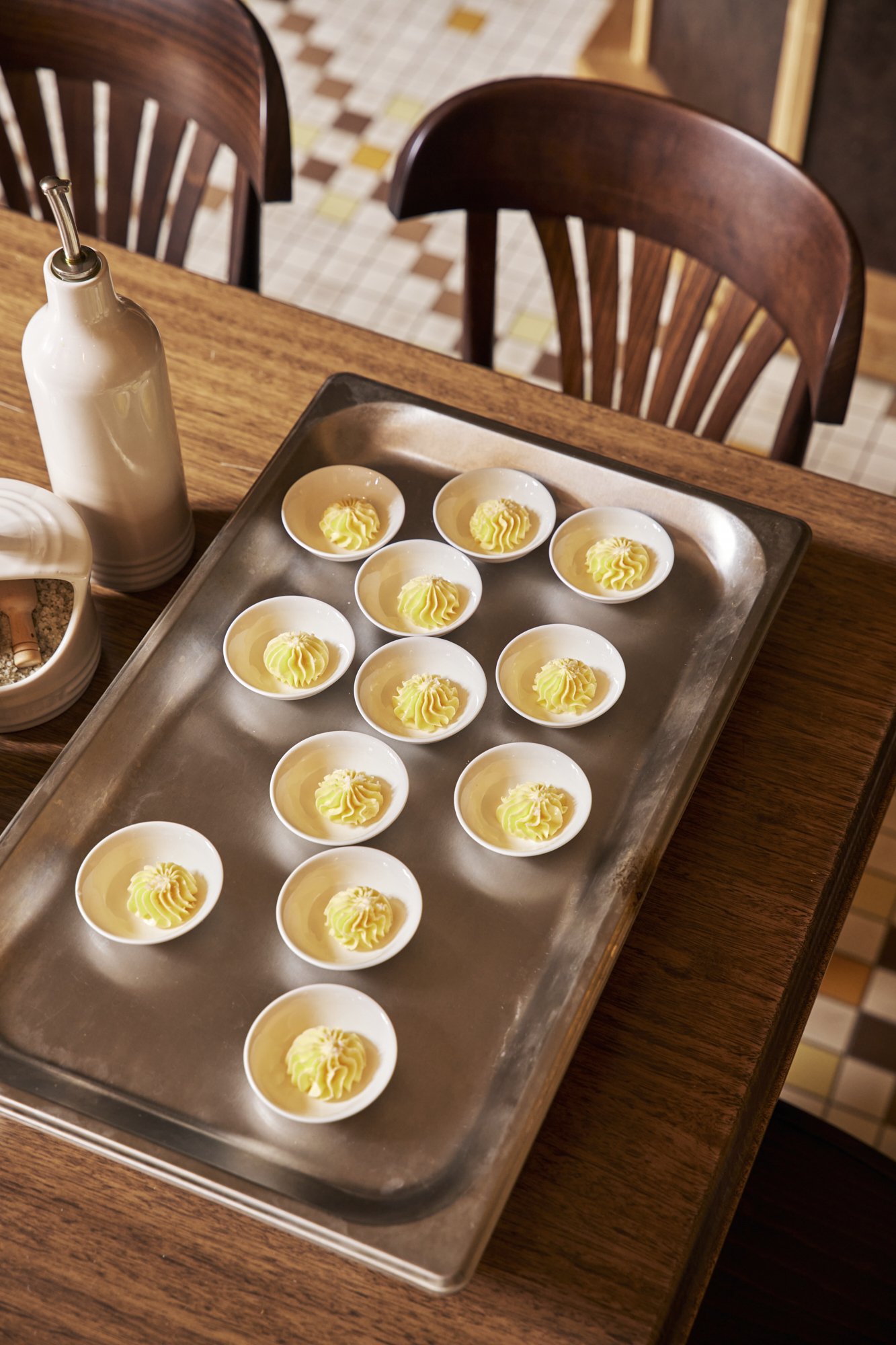 Small white dishes with yellow and green swirled butter curls arranged on a metal tray on a wooden table in a restaurant.
