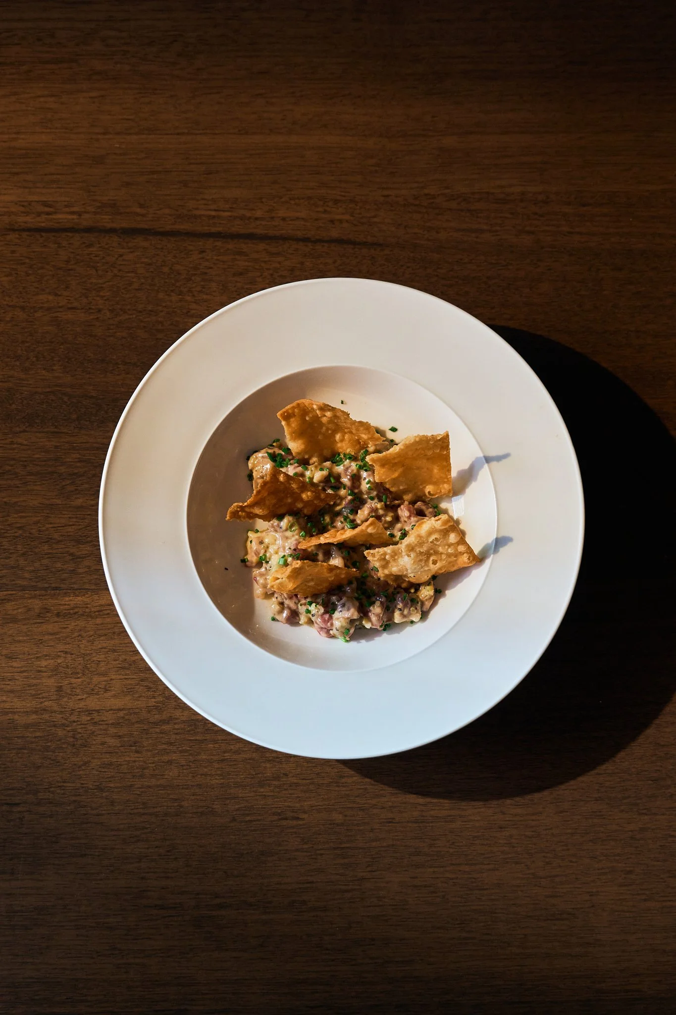A white bowl of tortilla chips, topped with chopped chives, served over a creamy dip or salad on a dark wooden table.