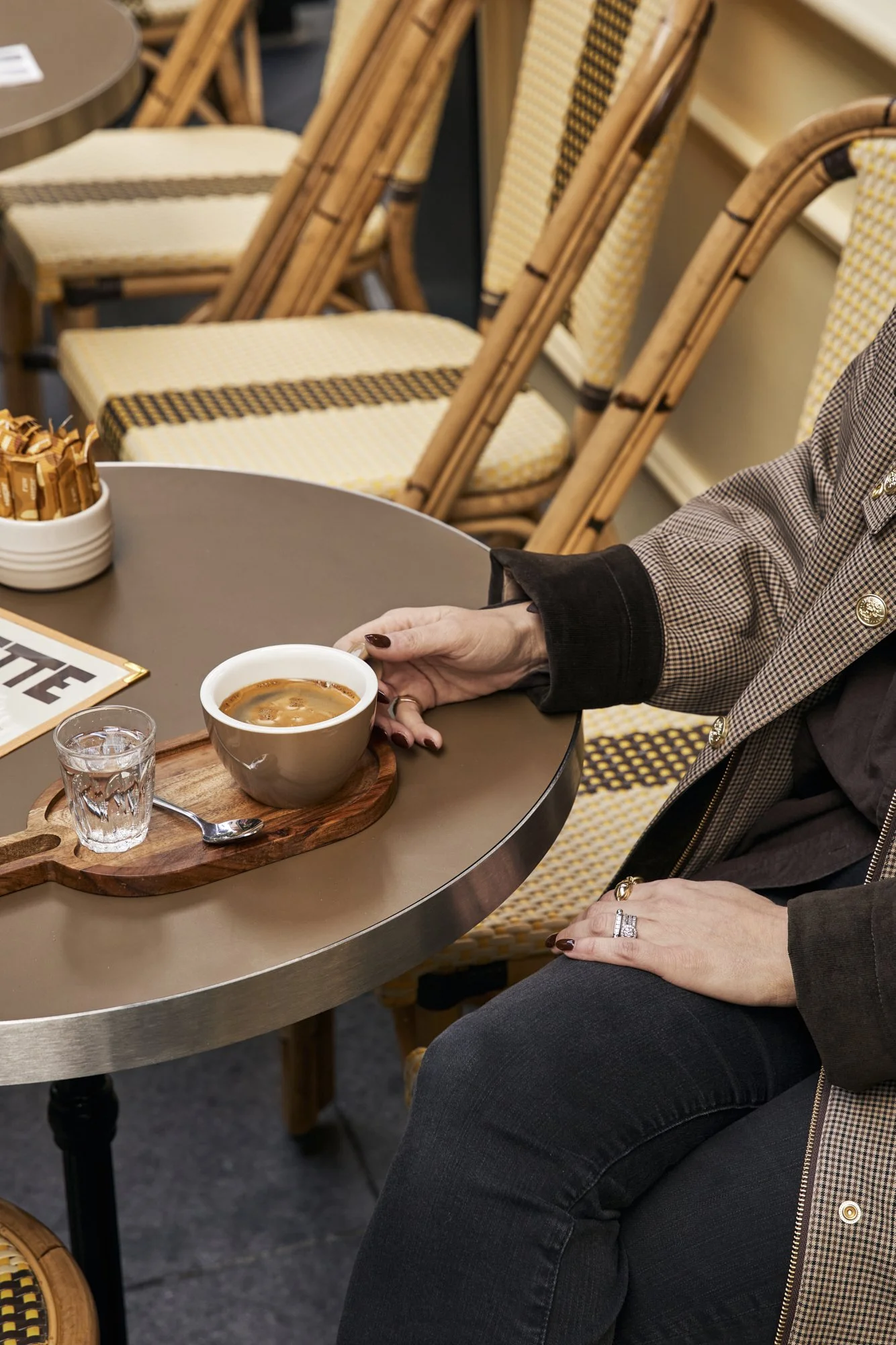 A person sitting at a table holding a cup of coffee, with a shot glass of water and a spoon on a wooden tray, in a cafe with empty woven chairs in the background.